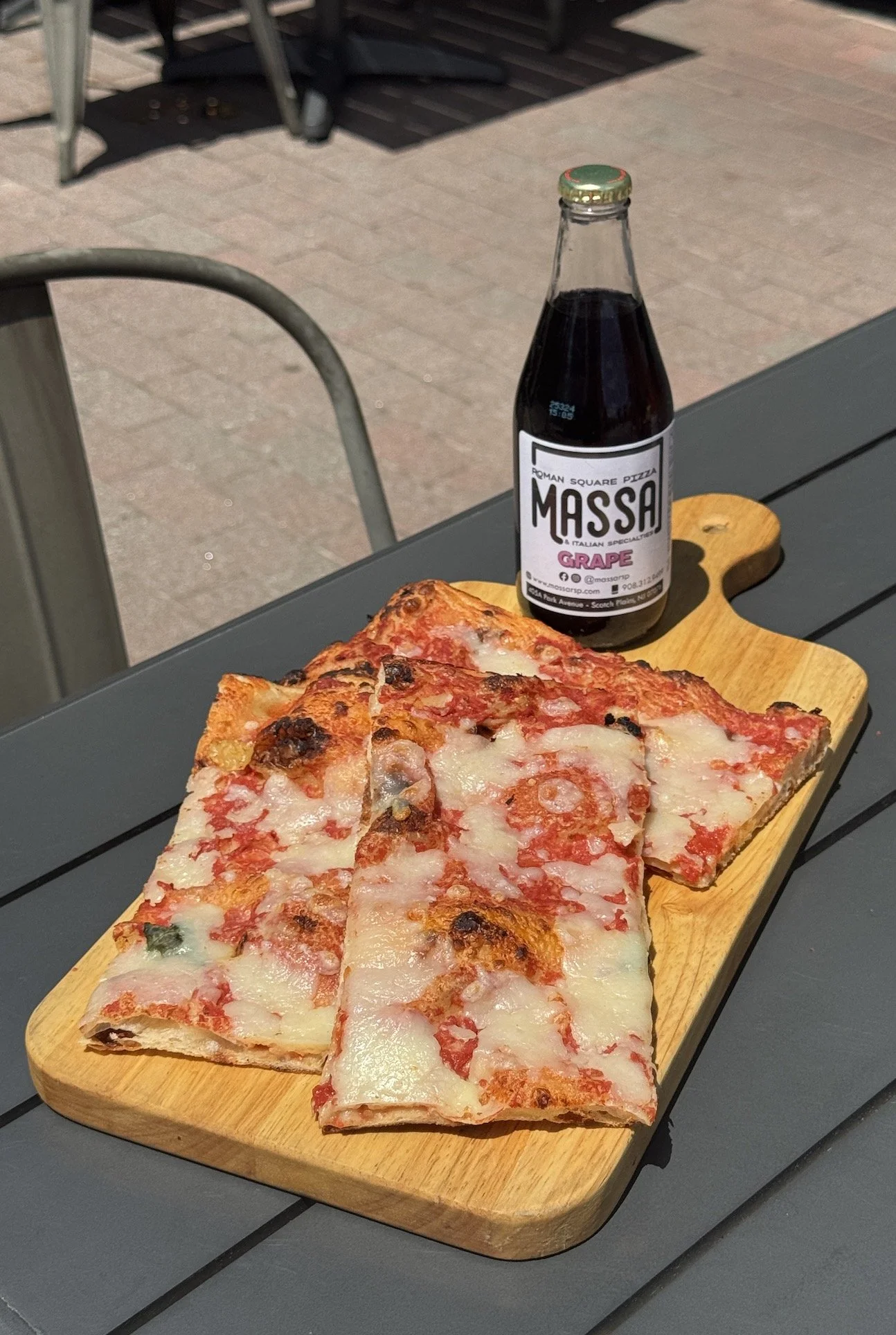 Slices of cheese and pepperoni pizza on a wooden serving board and a bottle of grape soda on an outdoor table.