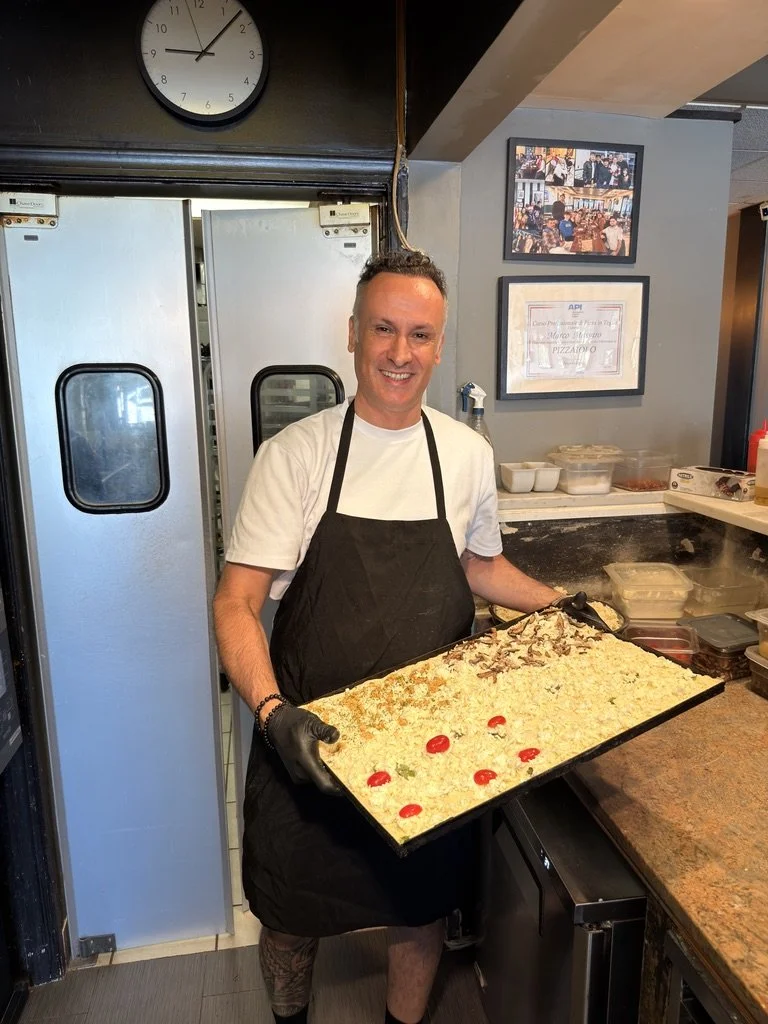 A man in a white t-shirt and black apron holds a large tray of cooked rice with cherry tomatoes and other toppings, standing inside a restaurant kitchen.