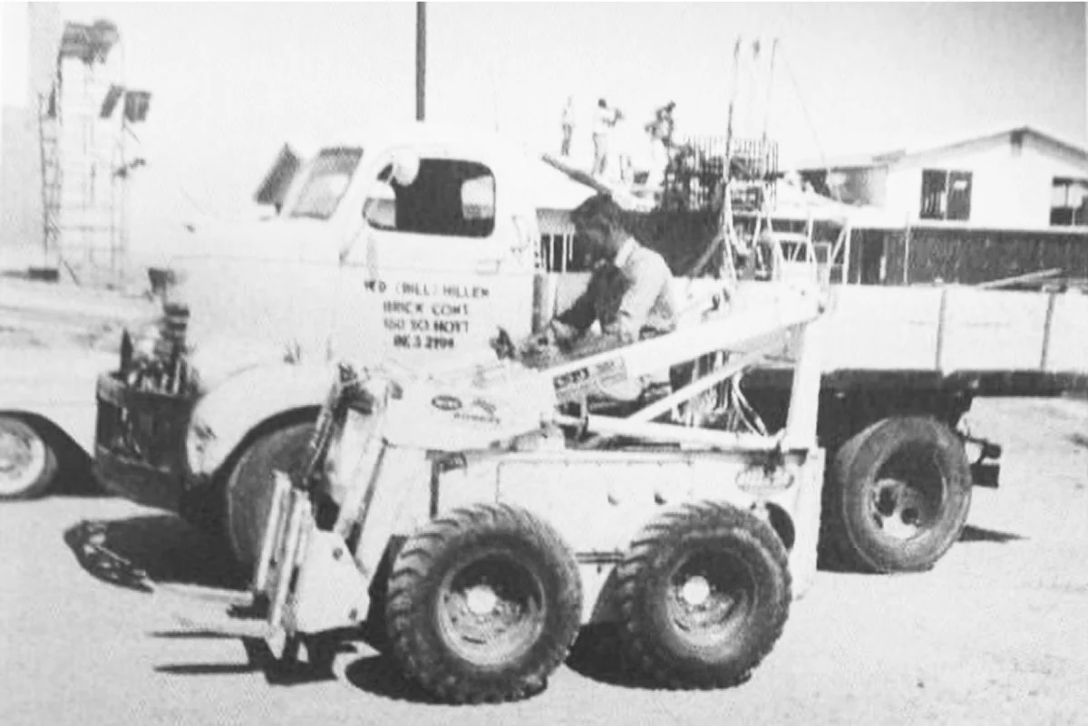Black and white photo of Will Hillen I operating a small front loader vehicle in a construction site with buildings and construction equipment in the background.