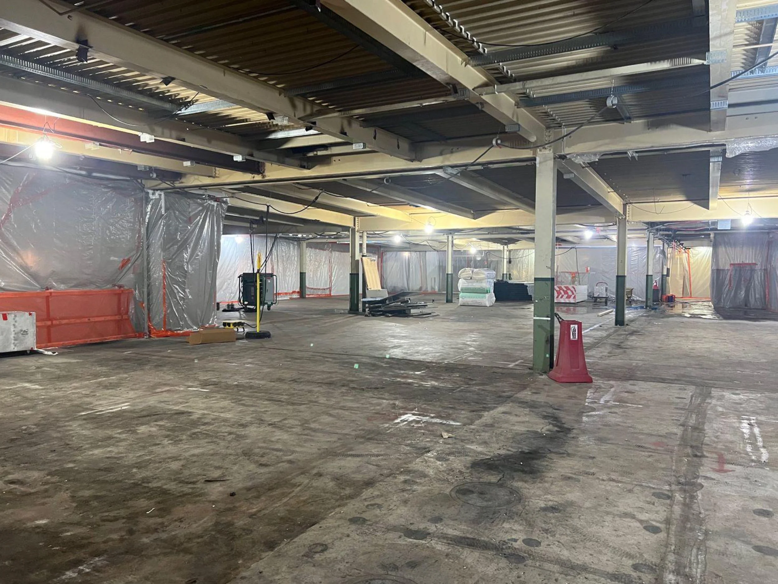 Construction site inside a building with exposed metal ceiling and support beams, plastic sheeting, construction materials, and equipment scattered on the unfinished concrete floor.