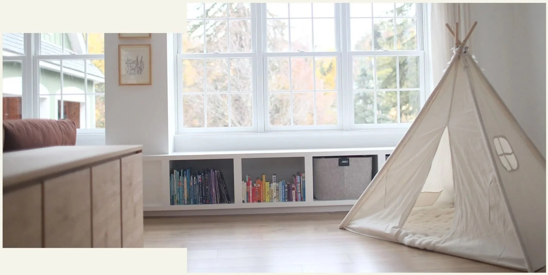 A cozy children's playroom with large windows, a white bookshelf filled with colorful books, a teepee tent, and a wooden piano to the left.