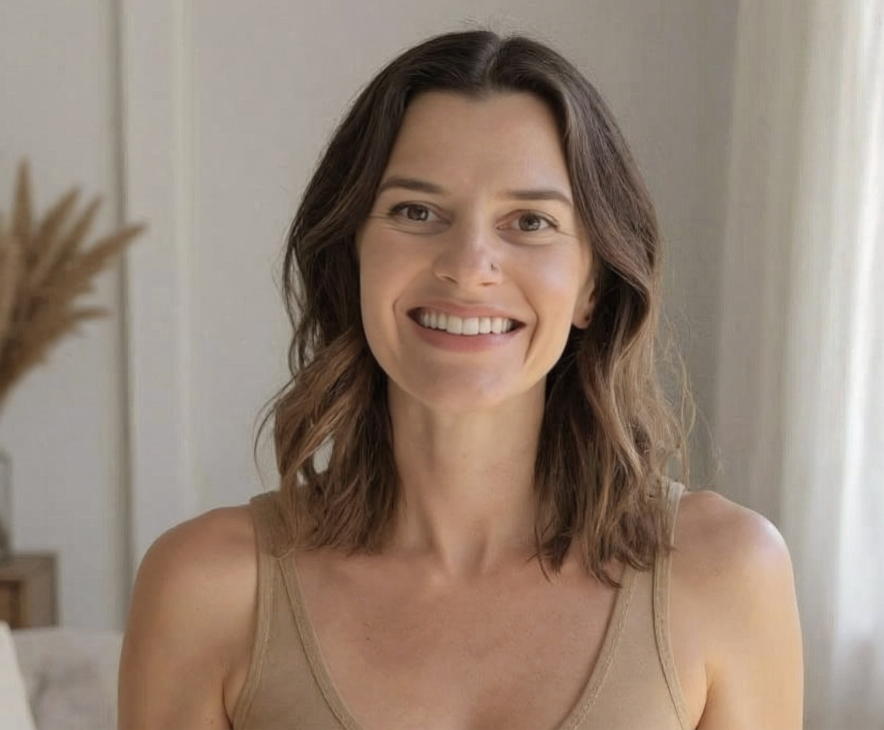 A woman with shoulder-length wavy brown hair and a nose piercing, smiling while looking at the camera, wearing a beige tank top. In a bright room with neutral decor in the background.