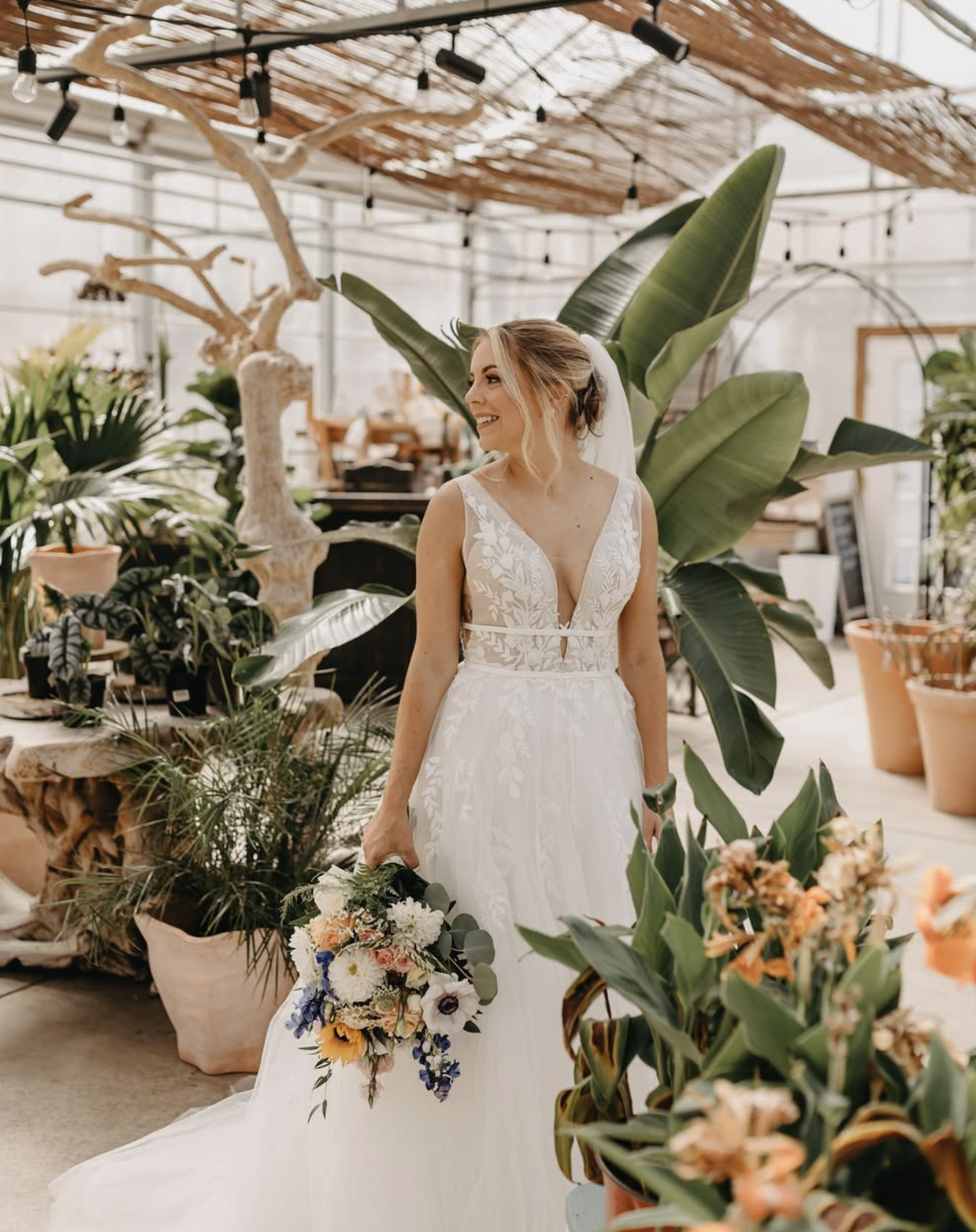 A woman in a white wedding dress holding a colorful flower bouquet inside a greenhouse filled with green plants and potted flowers.