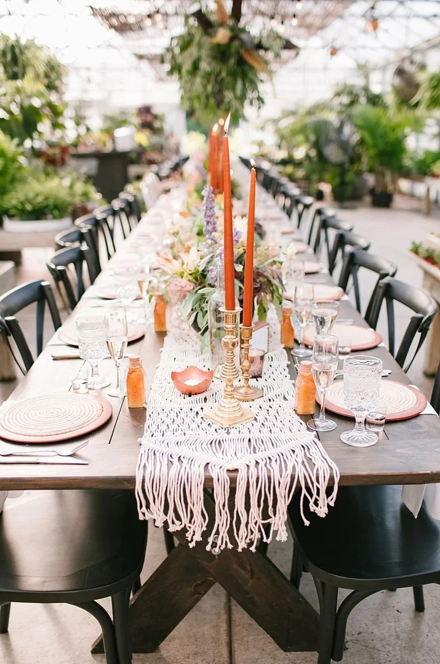 A long dining table set for a celebration with candles, flowers, place settings, and glassware inside a greenhouse or garden space.