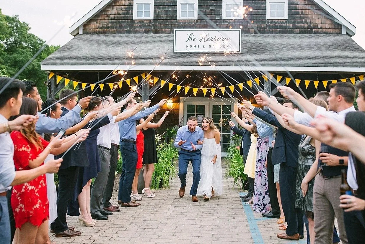 A bride and groom celebrating with guests in an outdoor wedding send-off, holding sparklers under a banner of yellow flags in front of a rustic building.