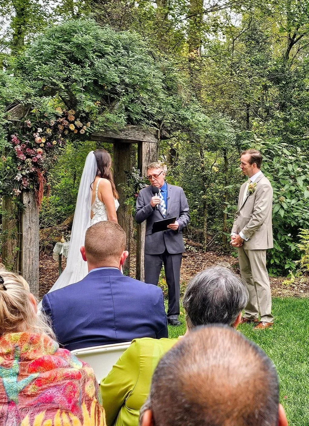 A wedding ceremony outdoors in a forest setting with a bride, groom, officiant, and guests seated. The bride is wearing a white wedding dress and veil, standing near a wooden arch decorated with flowers. The officiant is reading from a book, holding 