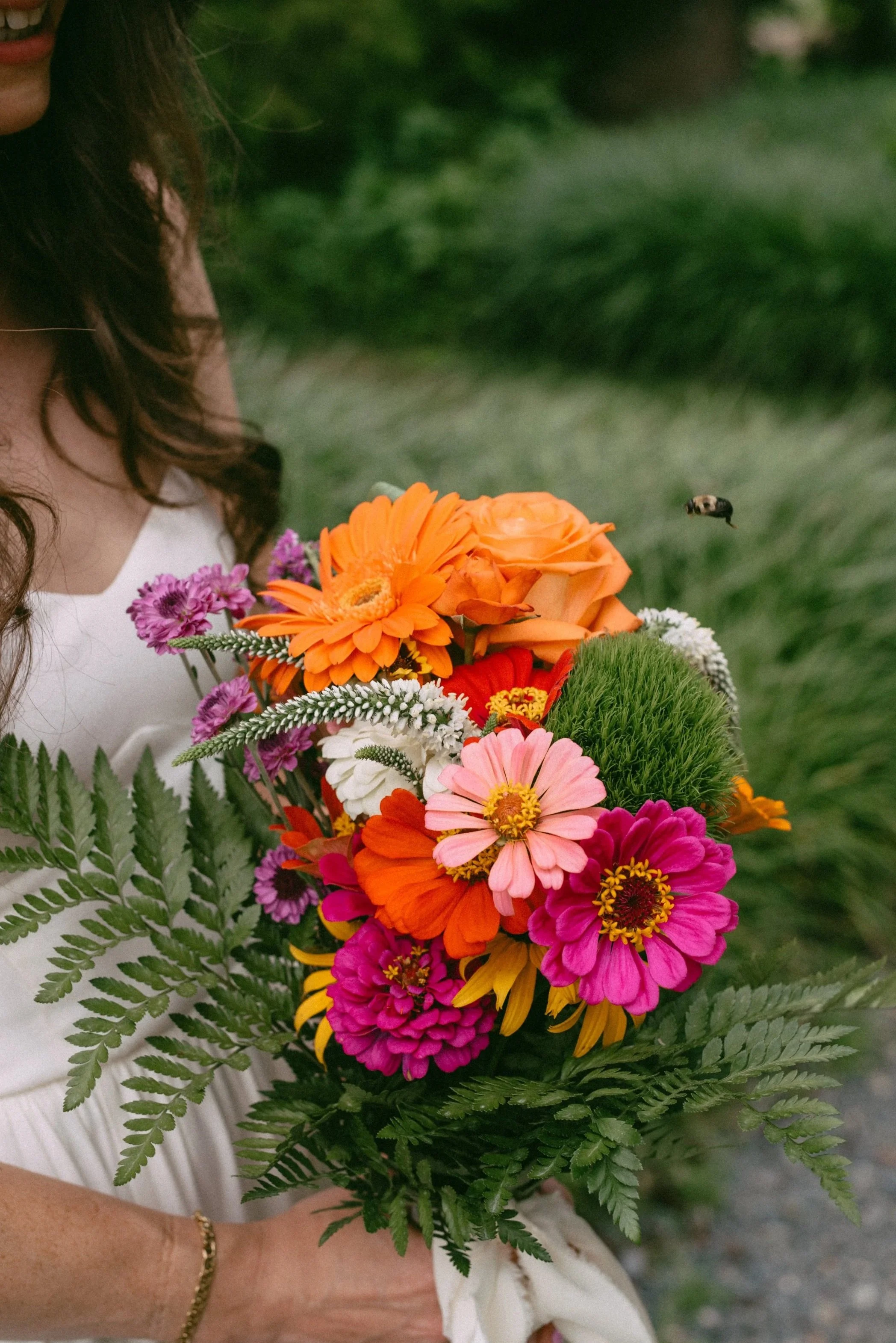 A woman holding a vibrant bouquet of multicolored flowers with green fern leaves, wearing a white dress and a gold bracelet on her wrist.