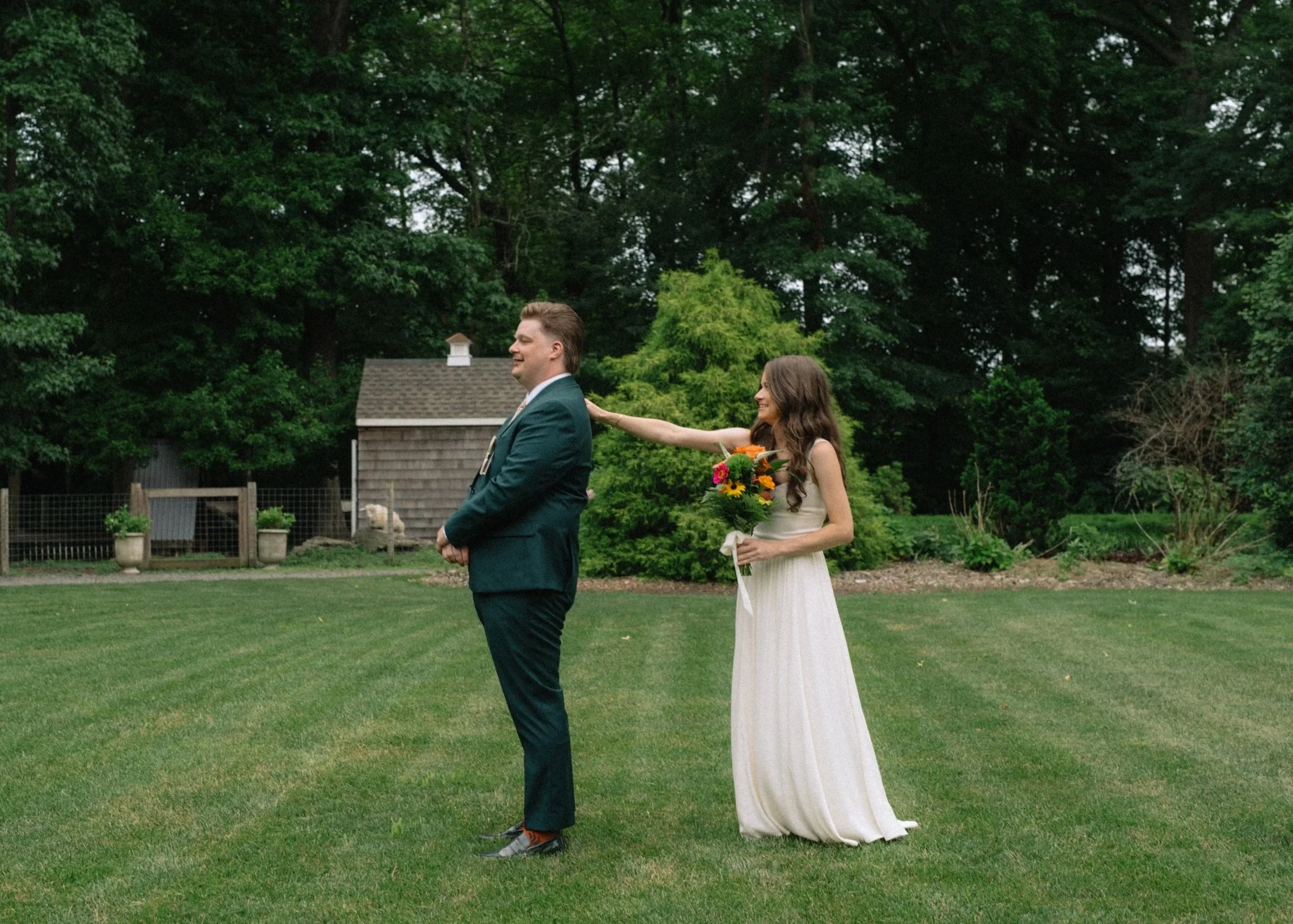 A bride and groom stand outdoors on a grassy area with trees in the background. The bride, holding a bouquet of sunflowers and orange flowers, is touching the groom's back. The groom is dressed in a suit and is smiling with his eyes closed.