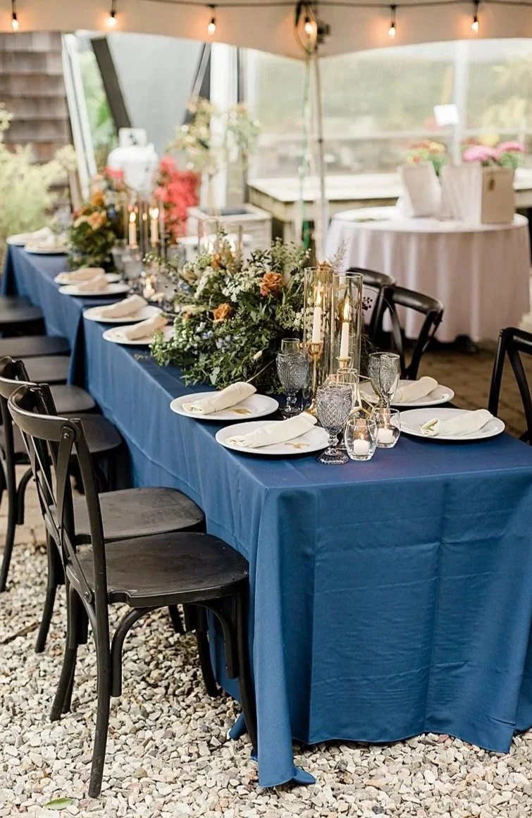 Long dining table with a dark blue tablecloth, decorated with a floral centerpiece, candle holders, and glassware, set for a formal event in a bright, enclosed outdoor space.