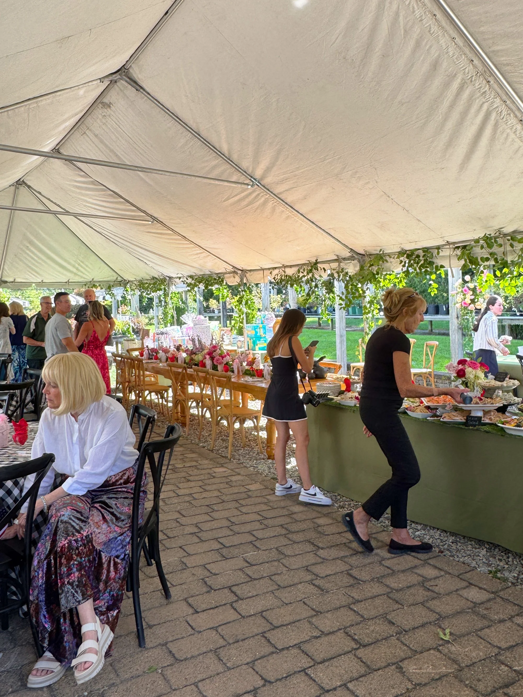 A group of people at an outdoor event under a large tent, with some people standing and others sitting at tables decorated with flowers, and a woman serving food at a buffet table.