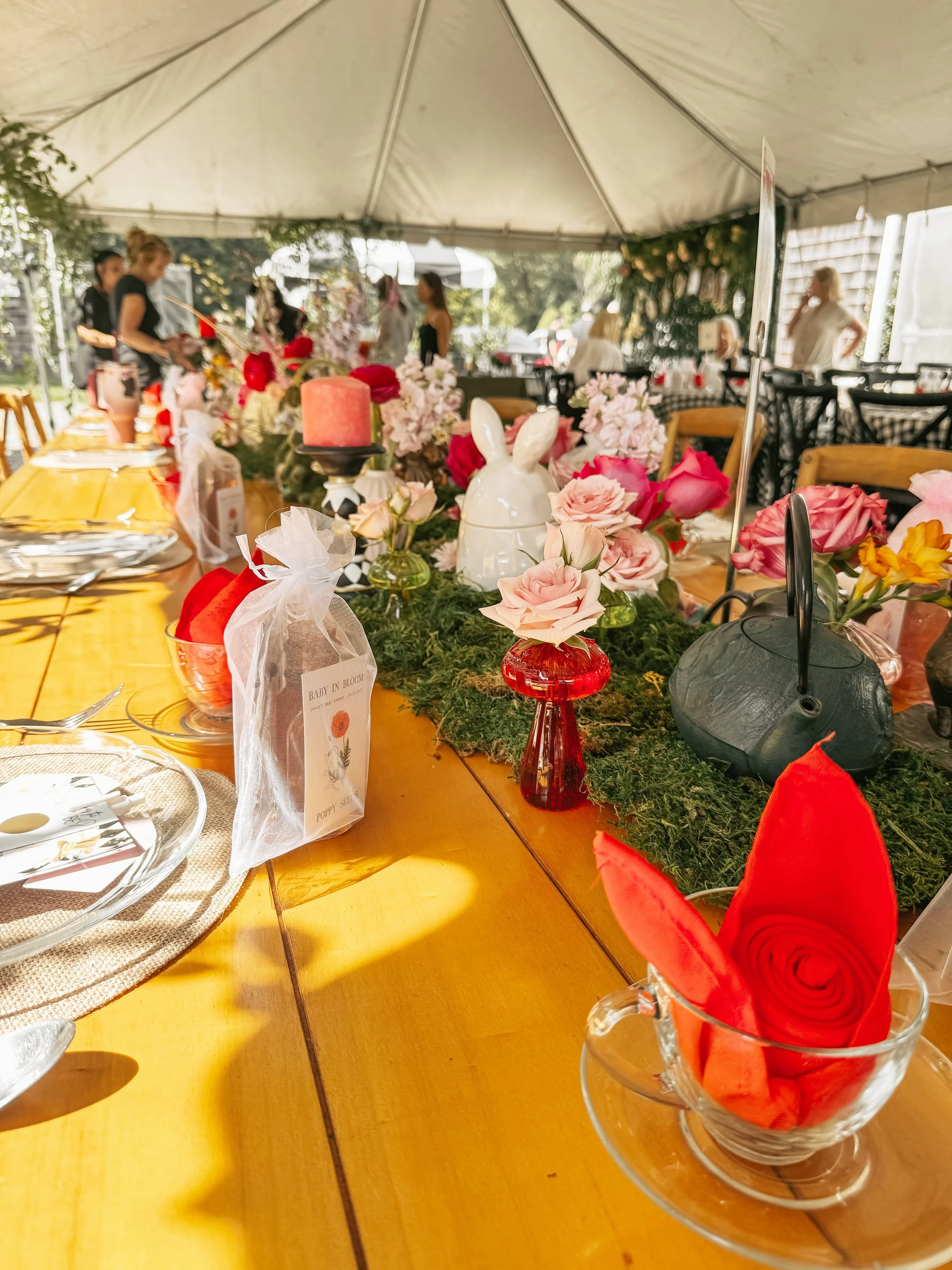 A decorated outdoor dining table with flowers, candles, and red cloth napkins under a white tent. People are visible in the background.