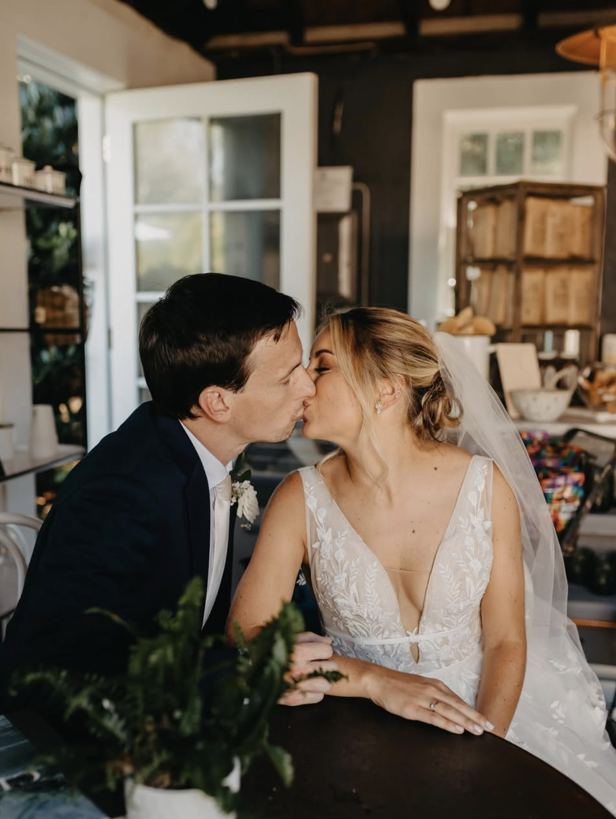 A bride and groom are sharing a kiss inside a cozy, rustic room with open windows and shelves filled with books and decorative items.