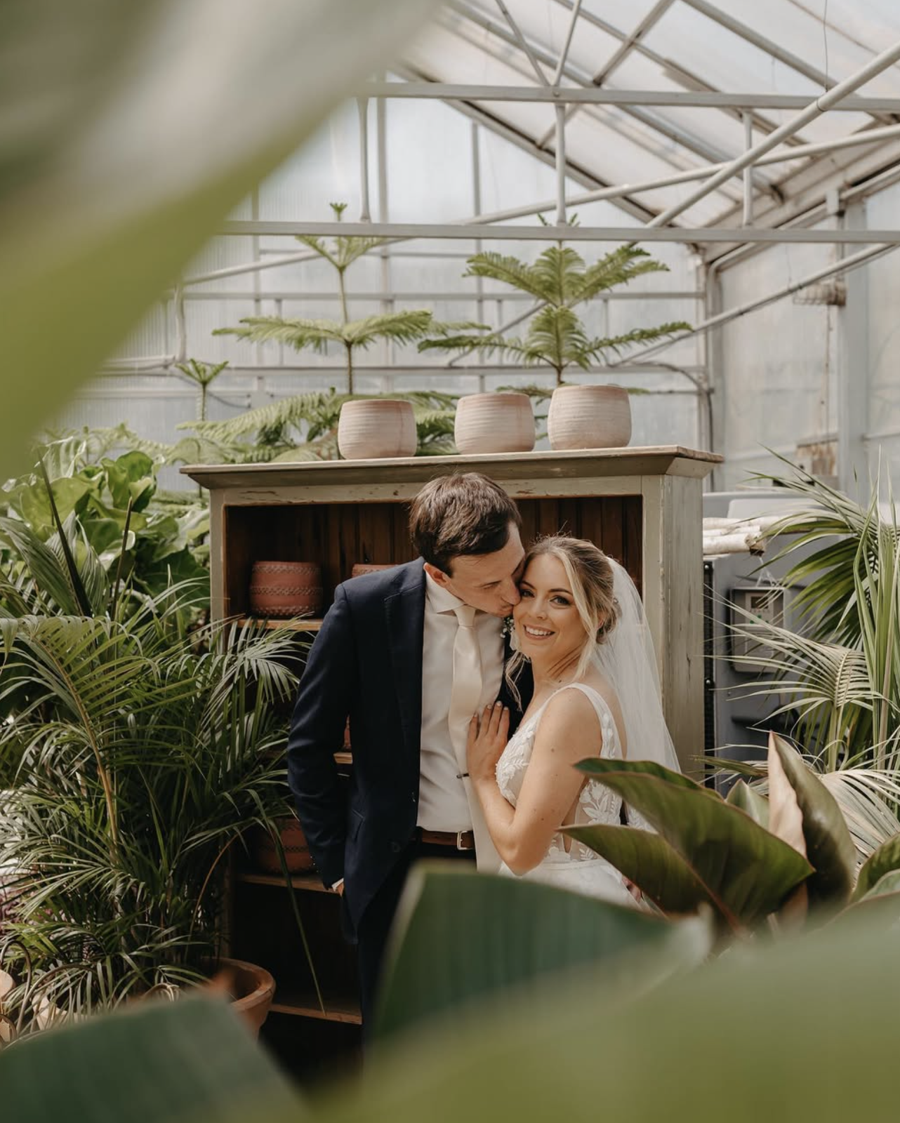 A newlywed couple in wedding attire embracing and smiling in a greenhouse surrounded by green plants.