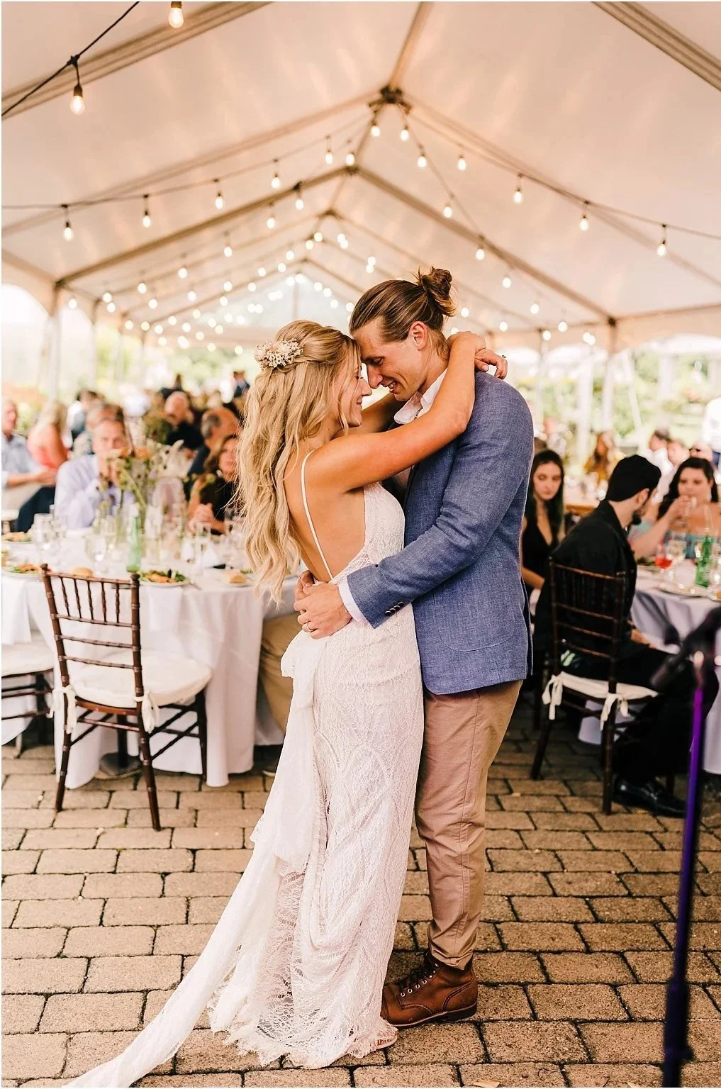 A bride and groom share a dance at their wedding reception inside a decorated tent, with guests seated at tables in the background.