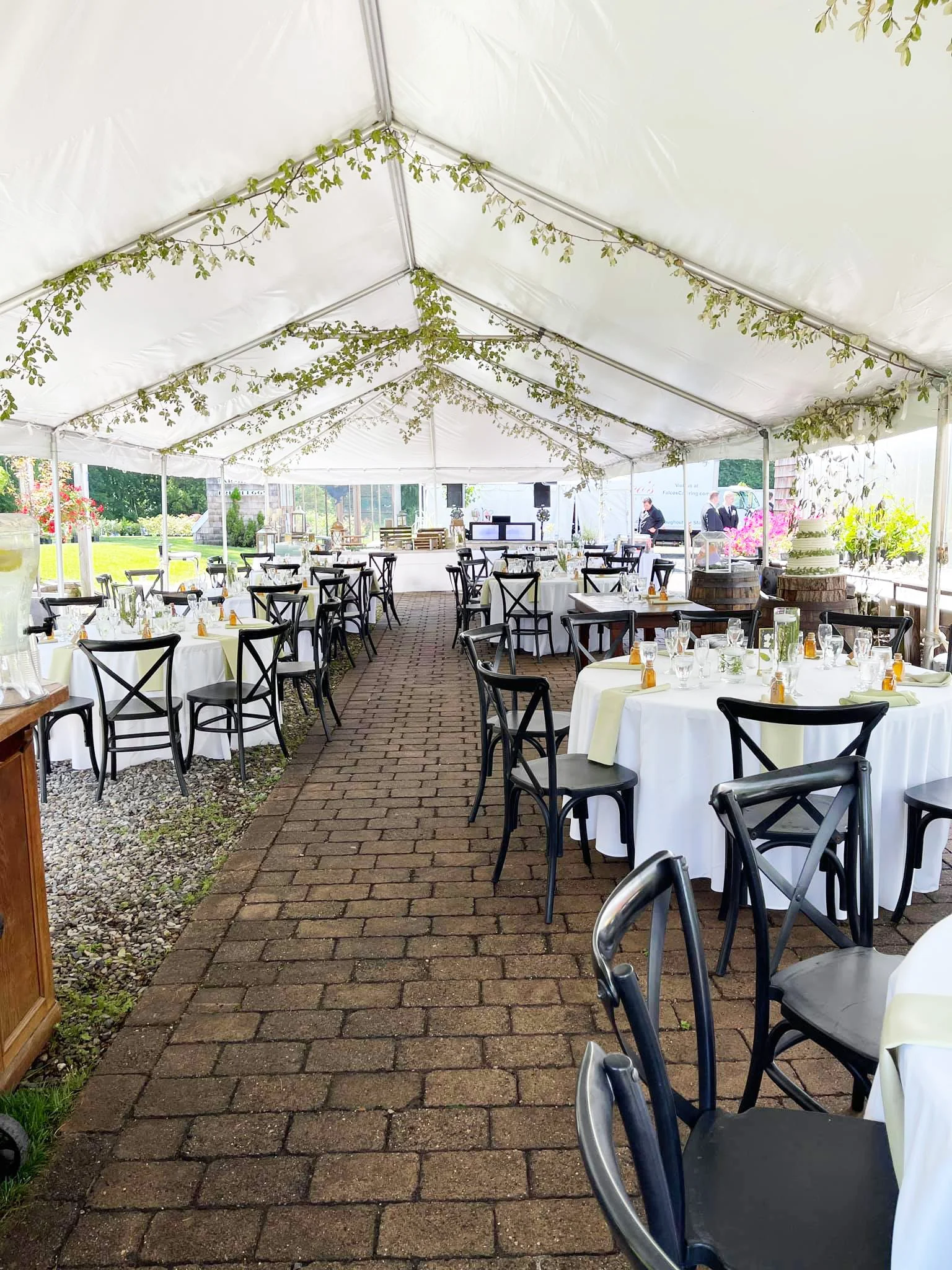 A white event tent set up outdoors with round tables covered in white tablecloths, black chairs, and a brick pathway. The tent is decorated with greenery along the ceiling, and there is a cake on a barrel at the right side. People are visible in the 