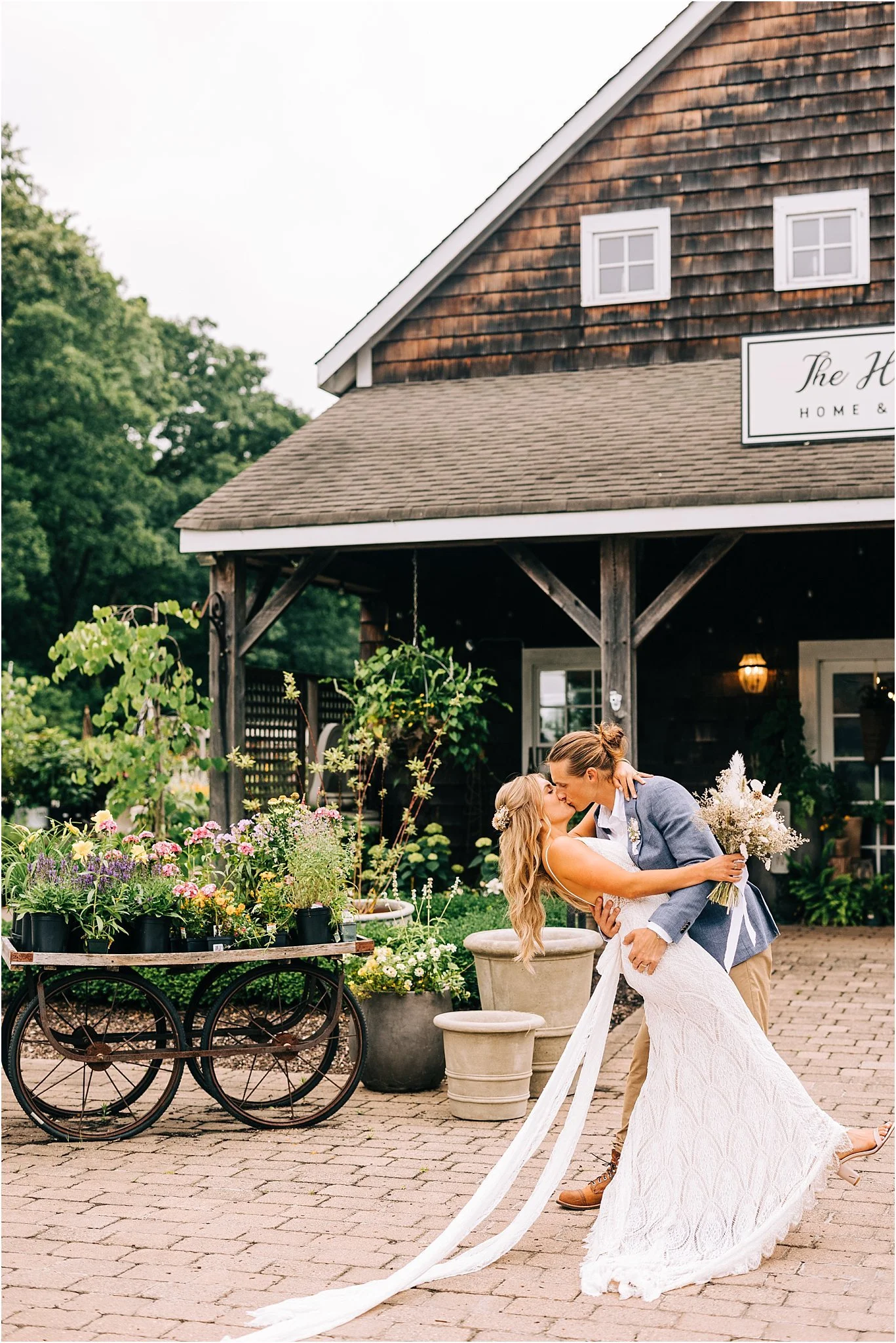 A bride and groom sharing a kiss during their outdoor wedding, with flowers and plants in the background and rustic barn-style building behind them.