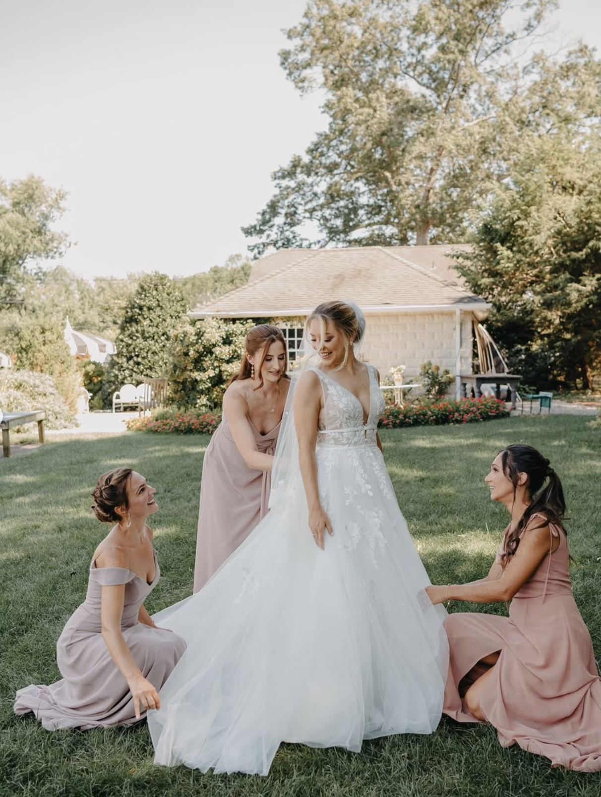 A bride in a wedding dress is being assisted by four women in bridesmaid dresses on a grassy outdoor area. They are all smiling, with a house, trees, and flowers in the background.