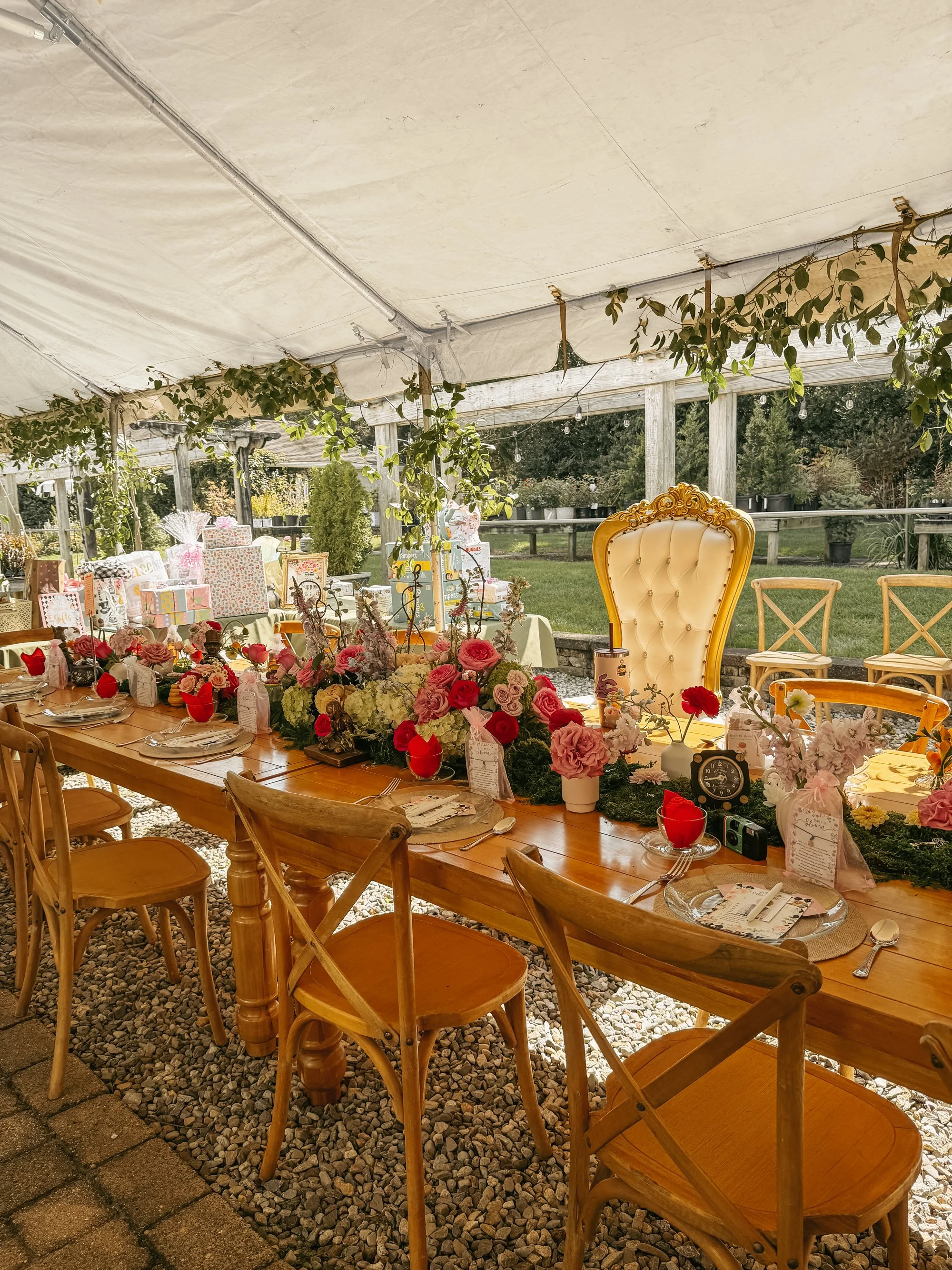 A long wooden table decorated with flowers, candles, and place settings, set outdoors under a large white tent with string lights and greenery, surrounded by chairs and a grassy yard.