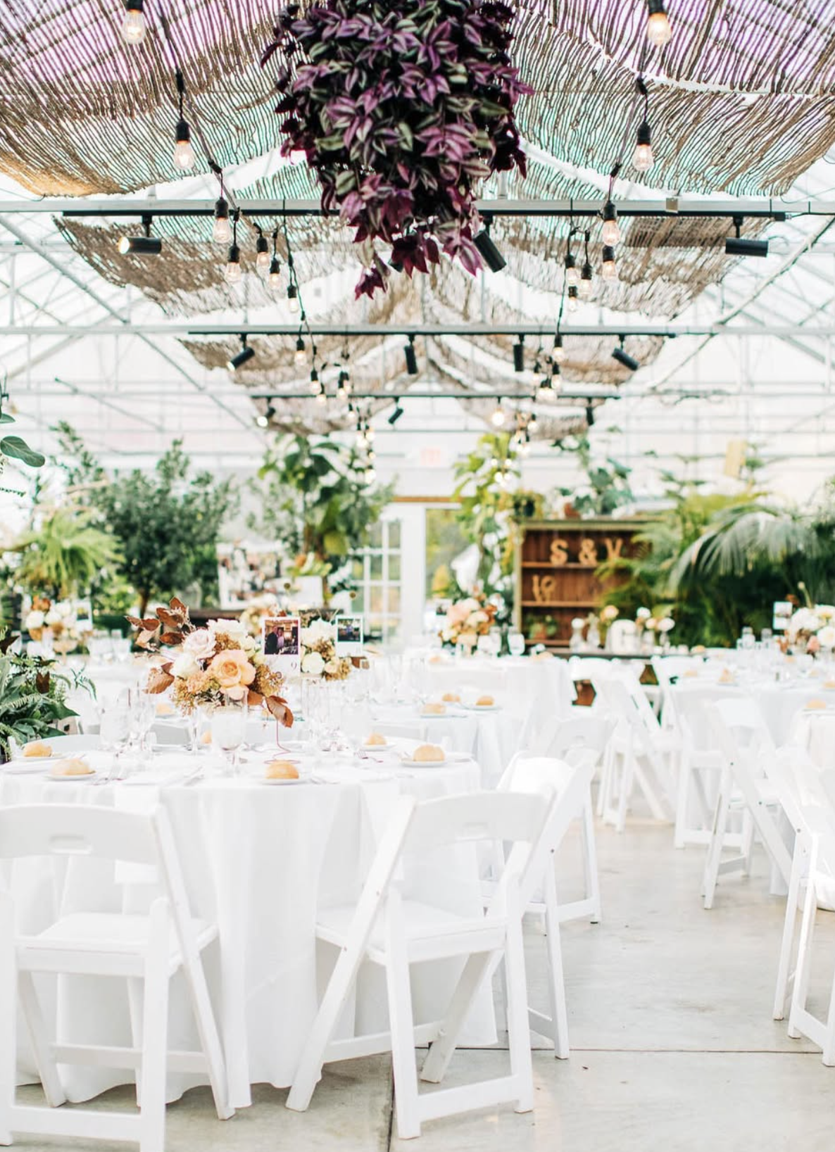 Wedding reception setup with round tables covered in white tablecloths, decorated with pink and white floral centerpieces, framed photos, and place settings. Green plants and trees decorate the space. The ceiling has hanging string lights and woven m