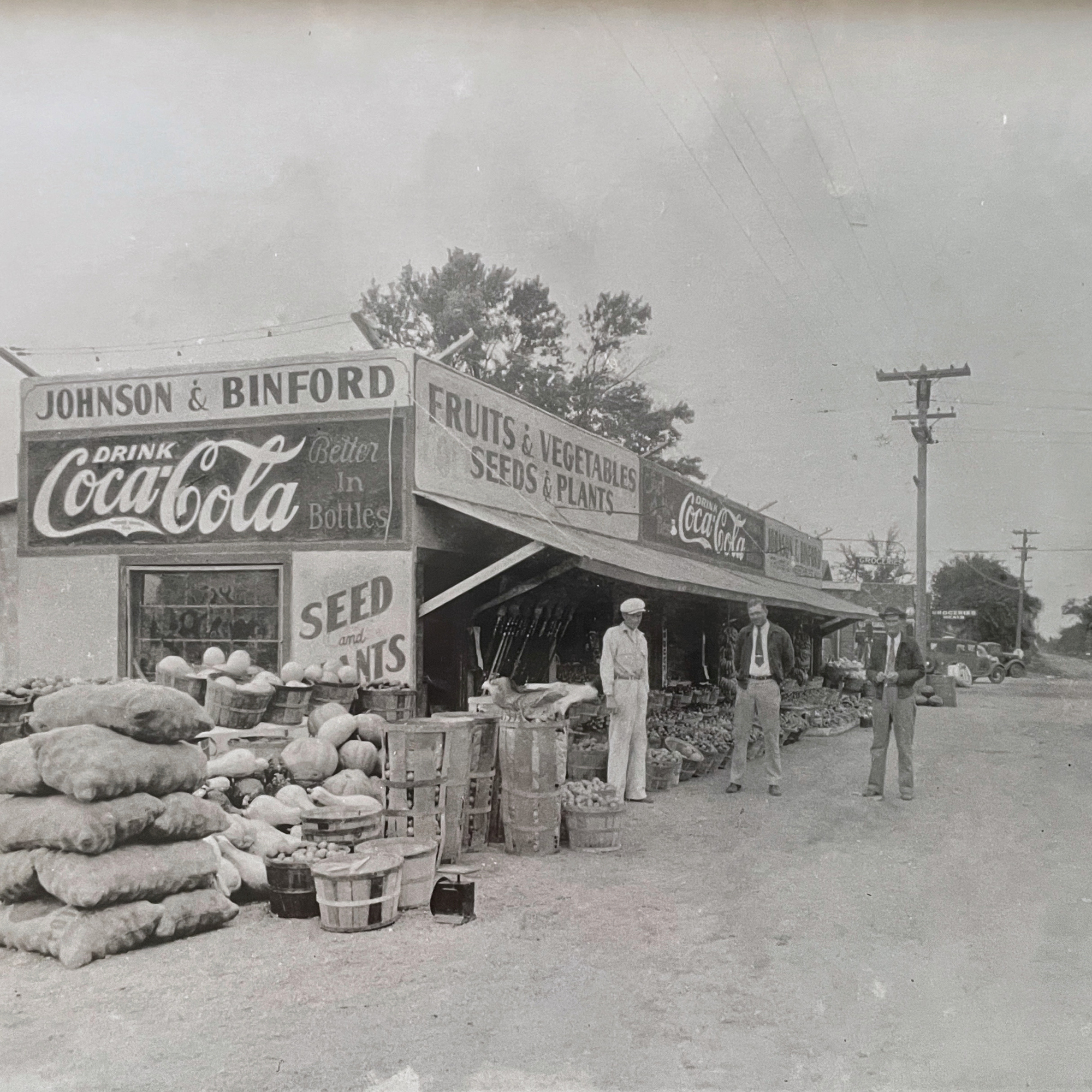 Black and white photo of a vintage farmers market stand with signs advertising Coca-Cola, fruits, vegetables, seeds, and plants, with three men standing outdoors in front of the stand, surrounded by baskets and sacks of produce, and vintage cars in the background.