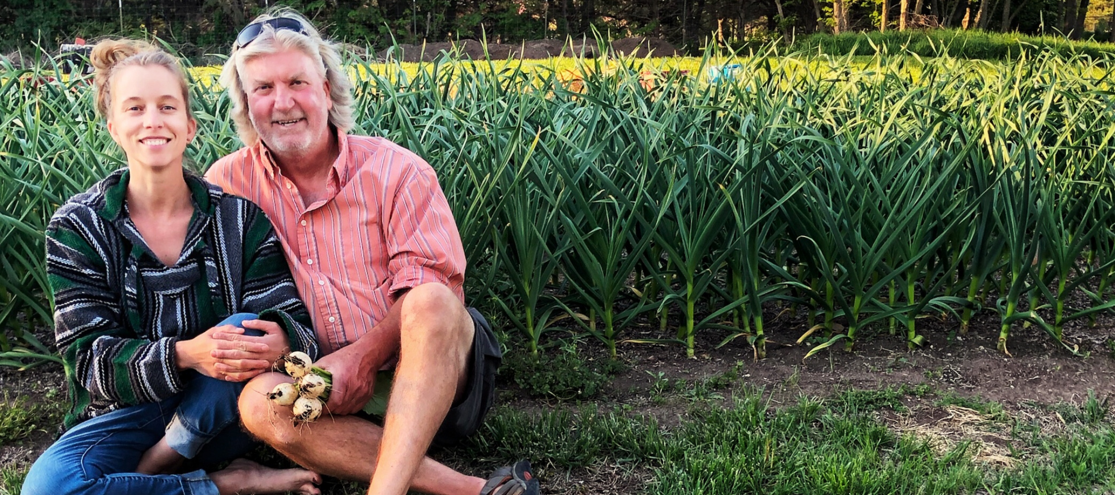 A smiling woman and man sitting in a field with tall garlic plants, holding a bunch of turnips.