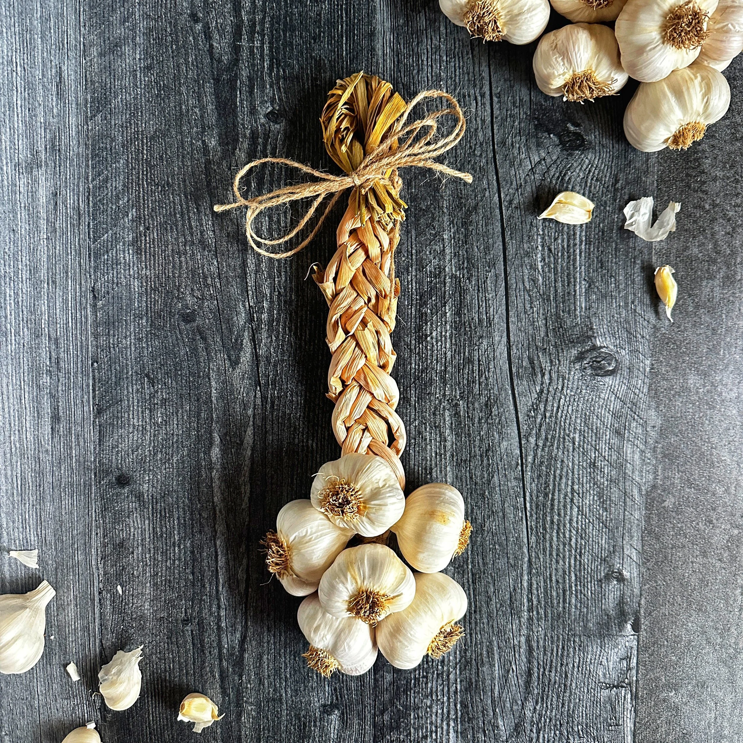 Centered flat-lay photograph of a handmade 6-bulb Mild French garlic braid resting vertically against a dark wood background.