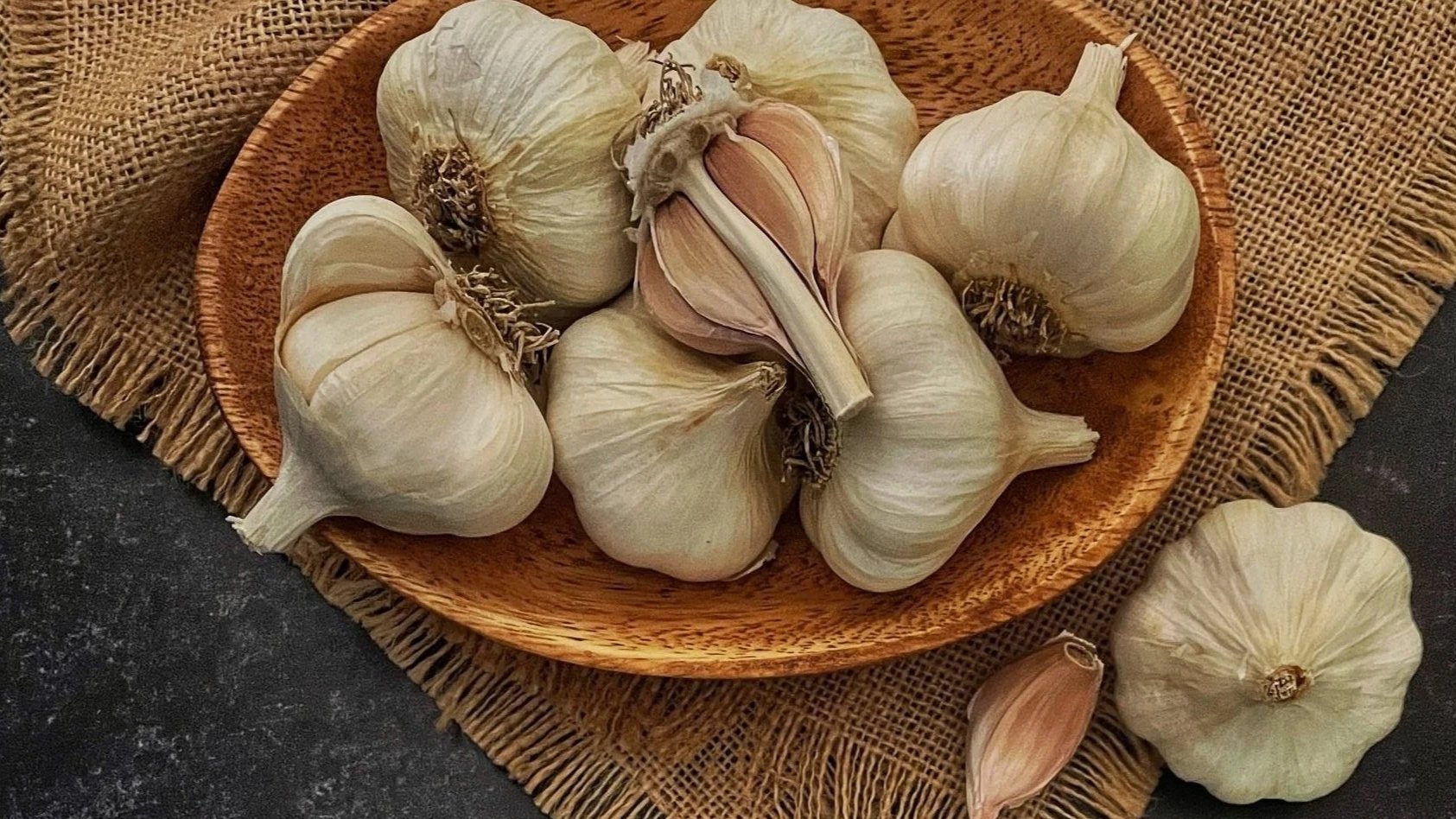 Garlic bulbs and cloves on a wooden bowl with a burlap cloth underneath.