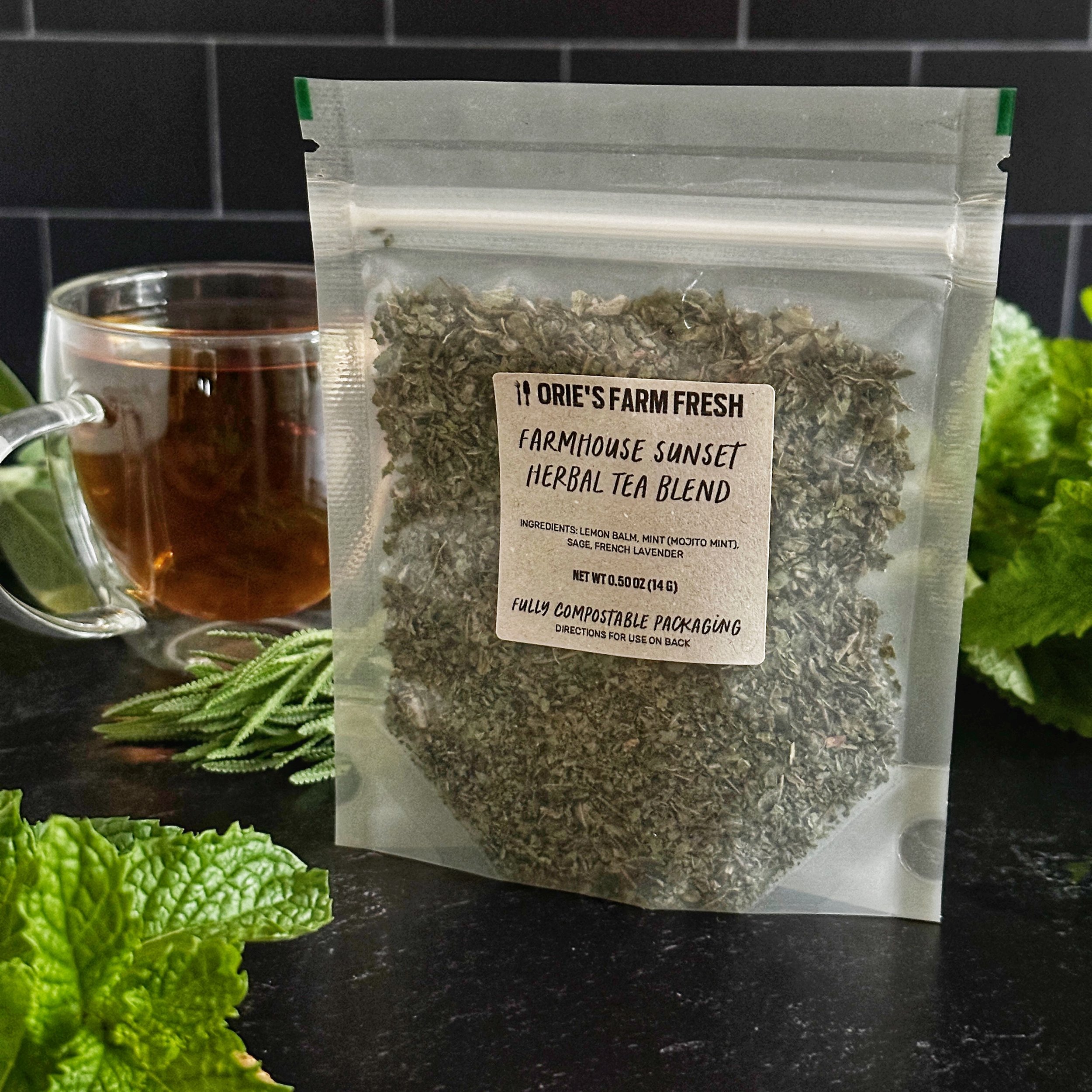 A golden-hued brewed cup of Farmhouse Sunset Herbal Tea Blend sits next to its compostable package with a black kitchen tile background.
