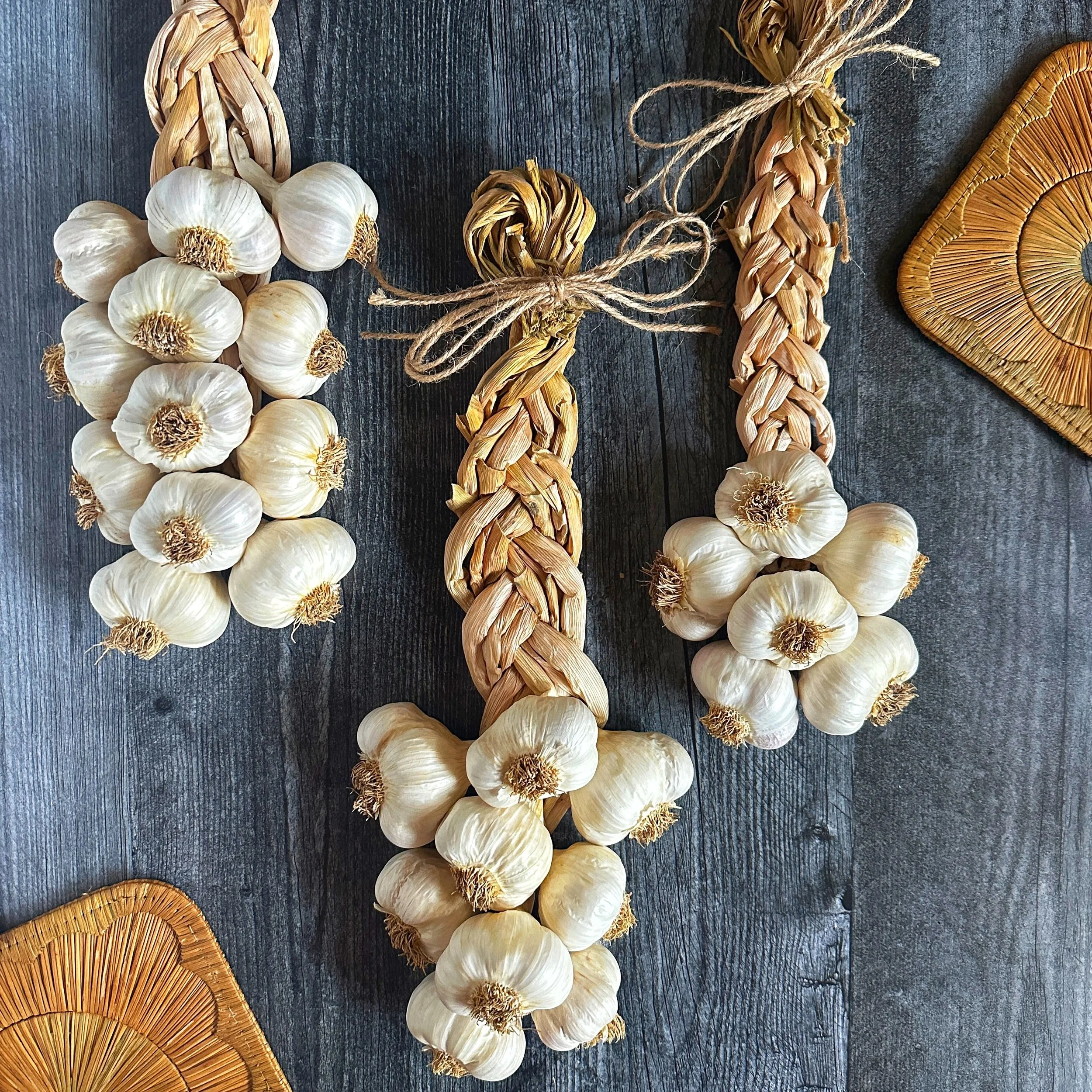 Flat-lay photograph of 6-bulb, 9-bulb, and 12-bulb handmade Mild French garlic braids arranged together on a dark wood background.