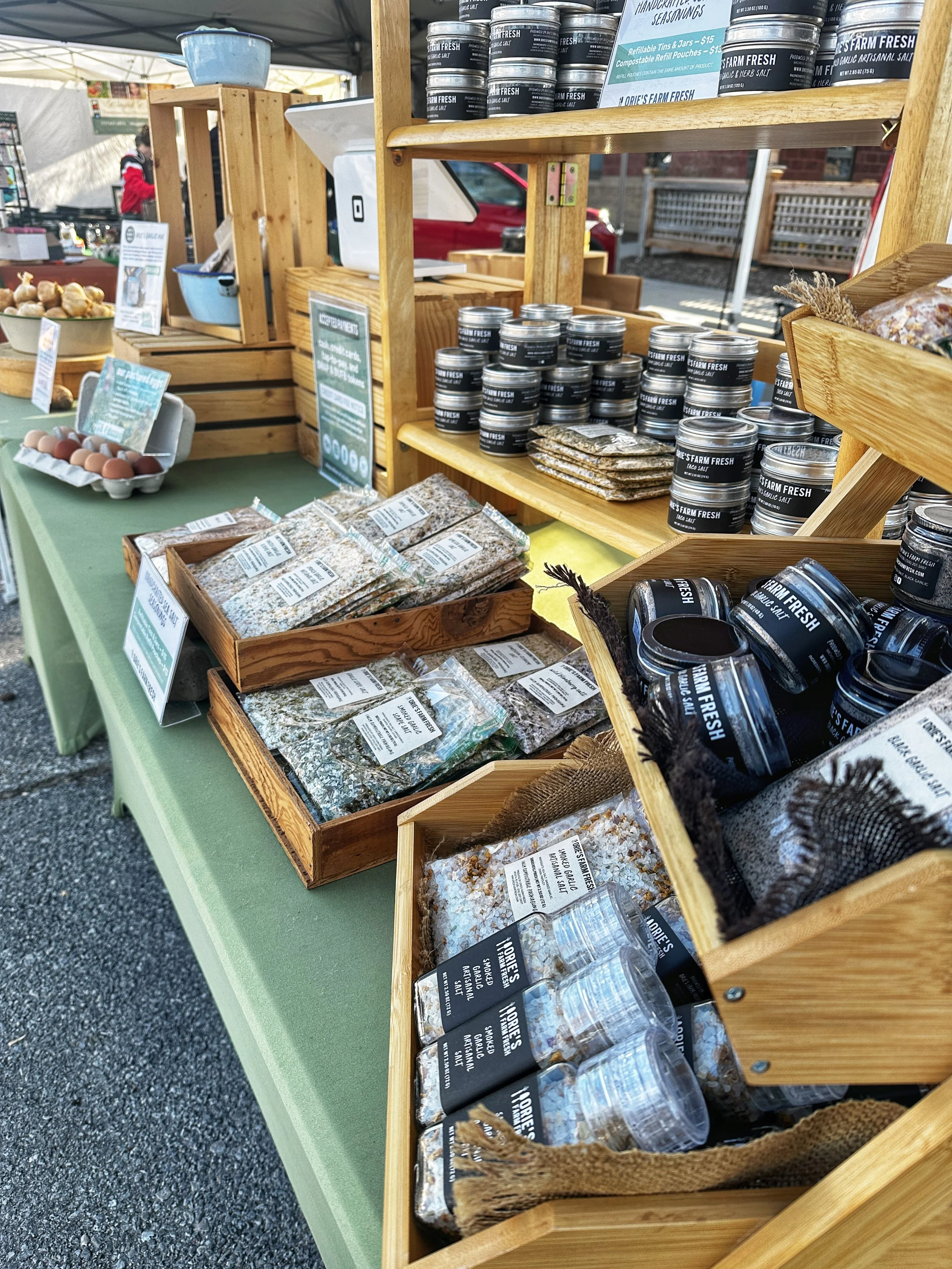 Market stand displaying various packaged food products, including seasonings and garlic powders, with a person in the background near a tent.