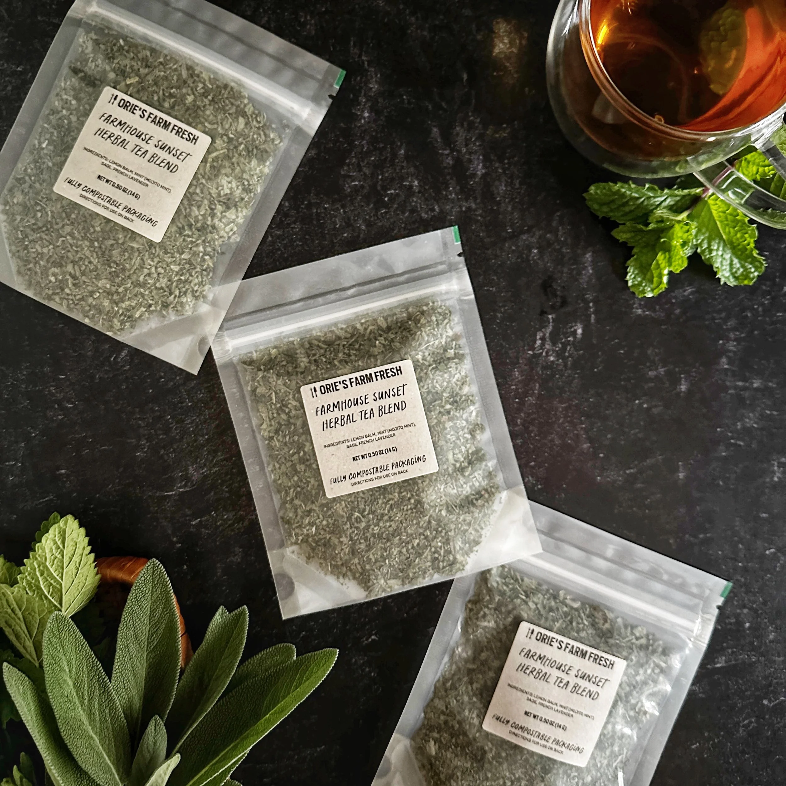 A flat-lay shot of 3 packages of Farmhouse Sunset Herbal Tea that features the golden-hued brewed cup of tea in the corner and freshly harvested garden herbs situated around the compostable packages.