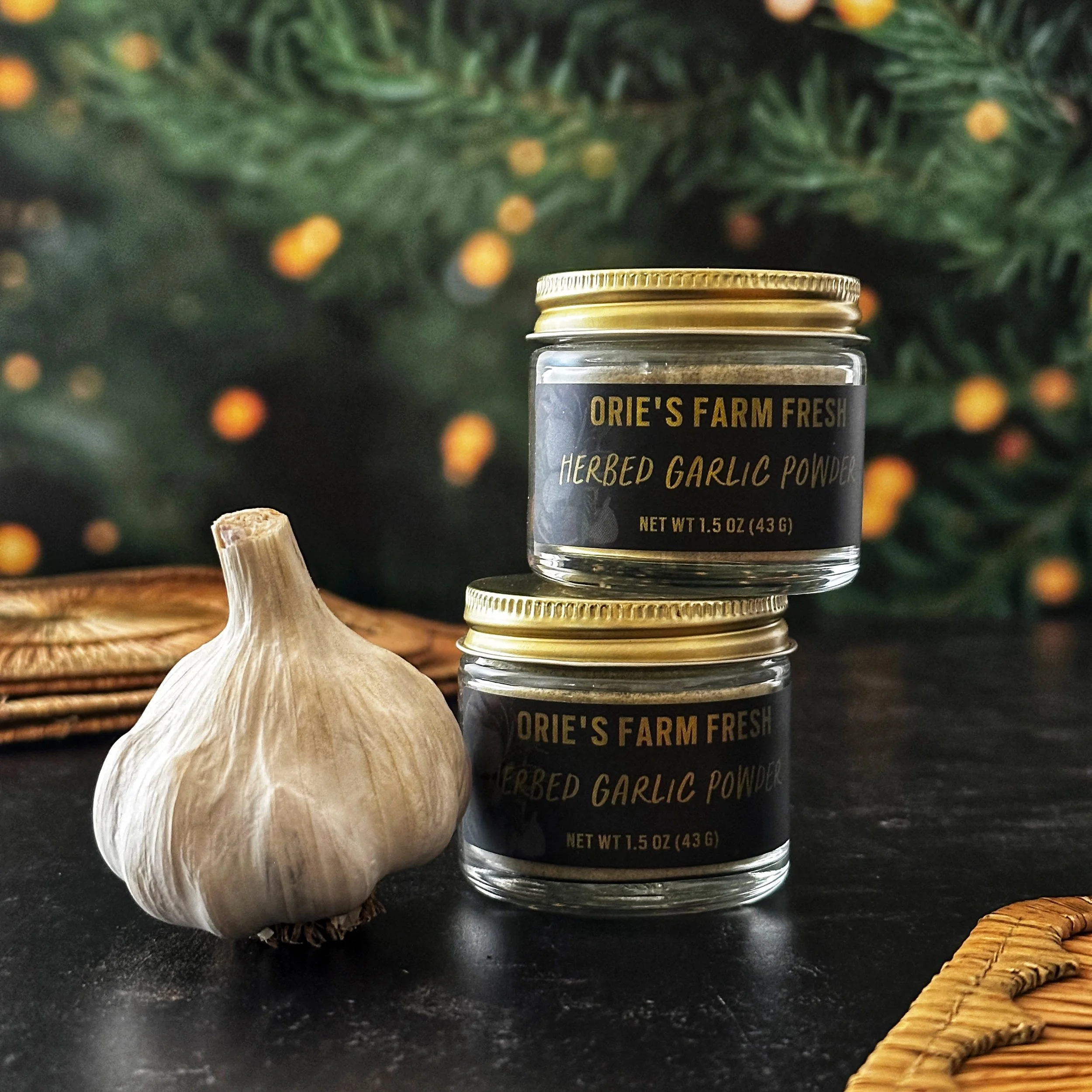 Herbed Garlic Powder jars look festive with their gold tins and a bulb of garlic next to them, all sitting on a slate surface in front of a lit Christmas tree.