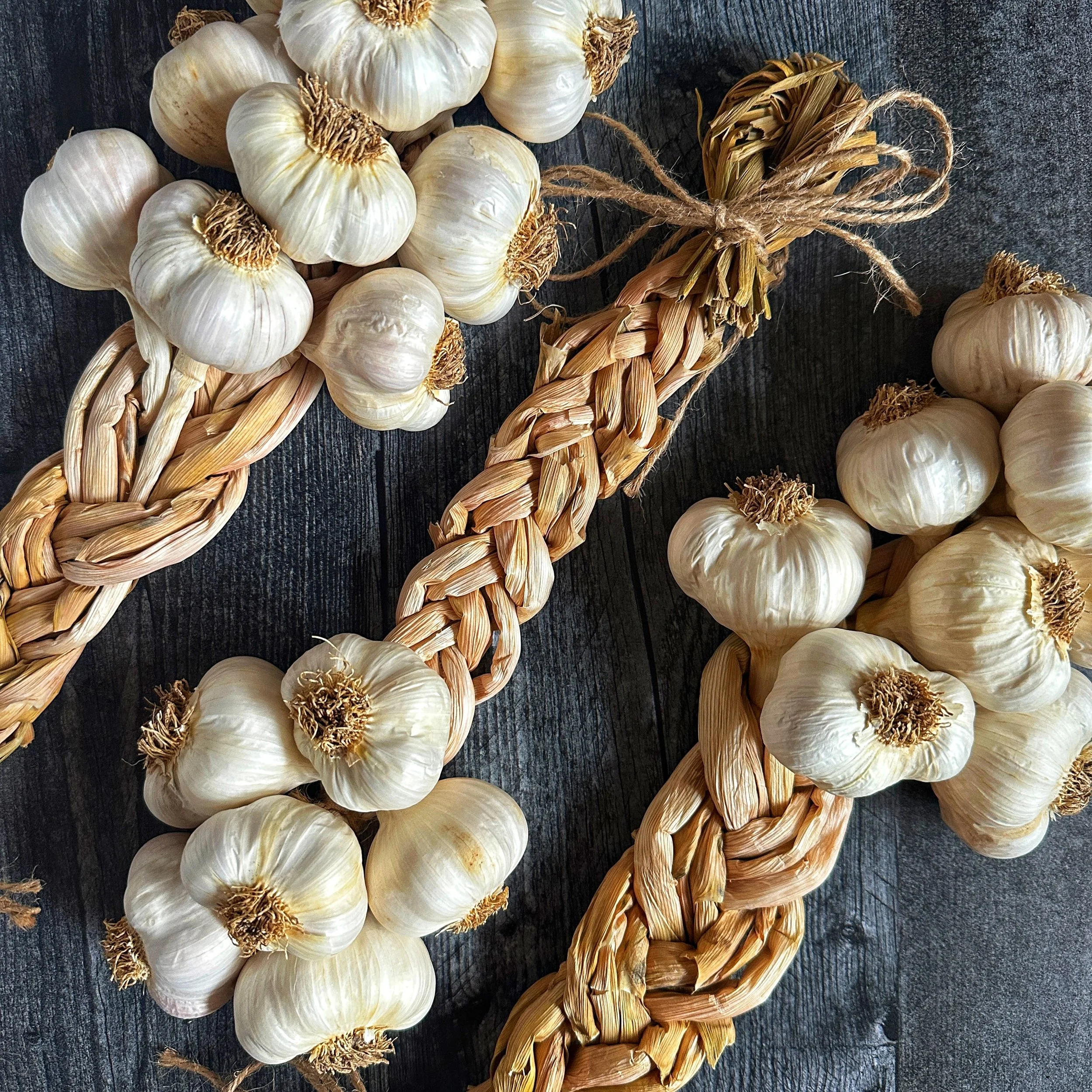 Diagonal close-up shot of three hand-woven Mild French garlic braids resting on a dark wood background, showing the woven stems and smooth bulb wrappers.