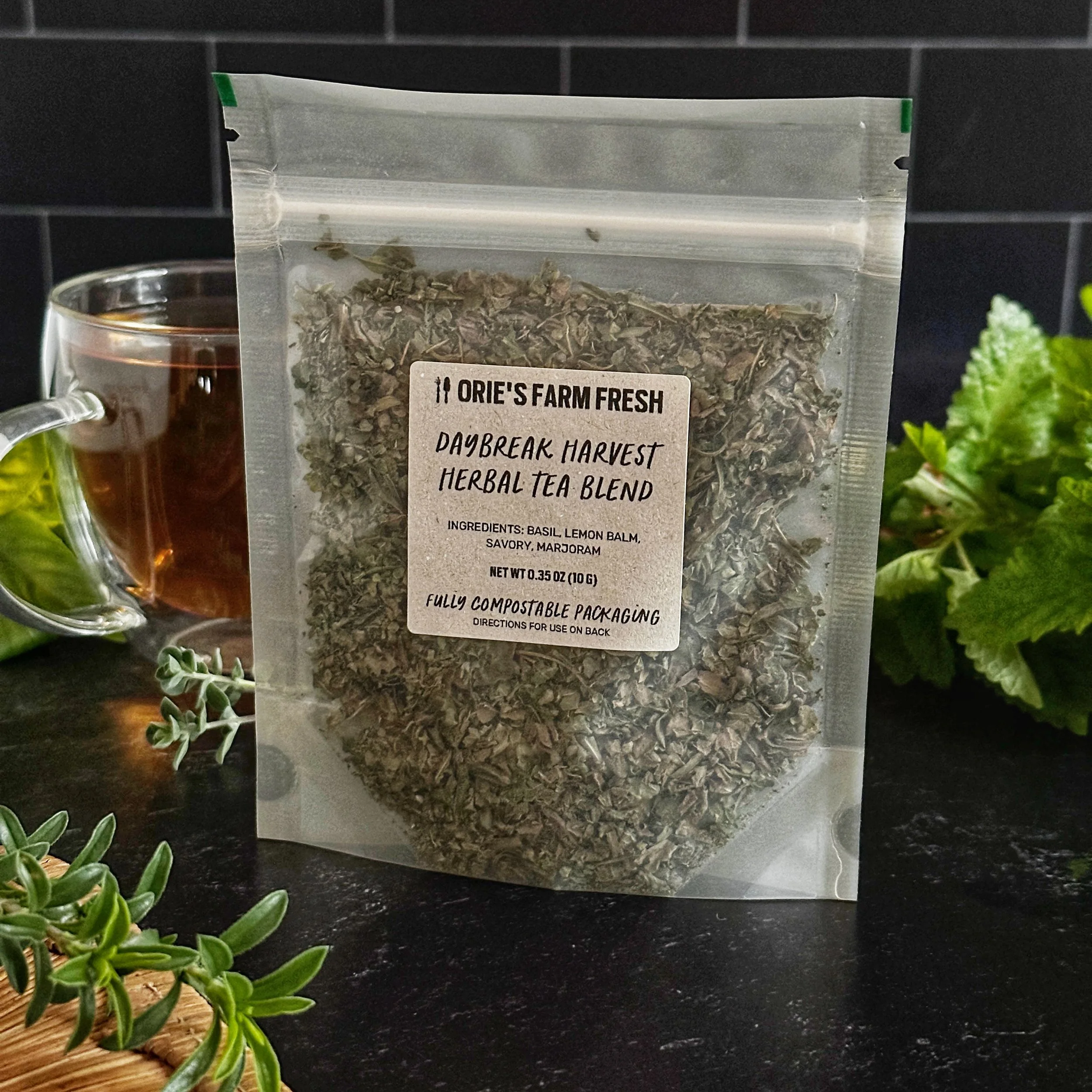 A fully compostable package of Daybreak Harvest Herbal Tea Blend sits centered on dark slate, with a clear mug of golden-hued tea sitting behind it, with fresh herbs in the foreground.