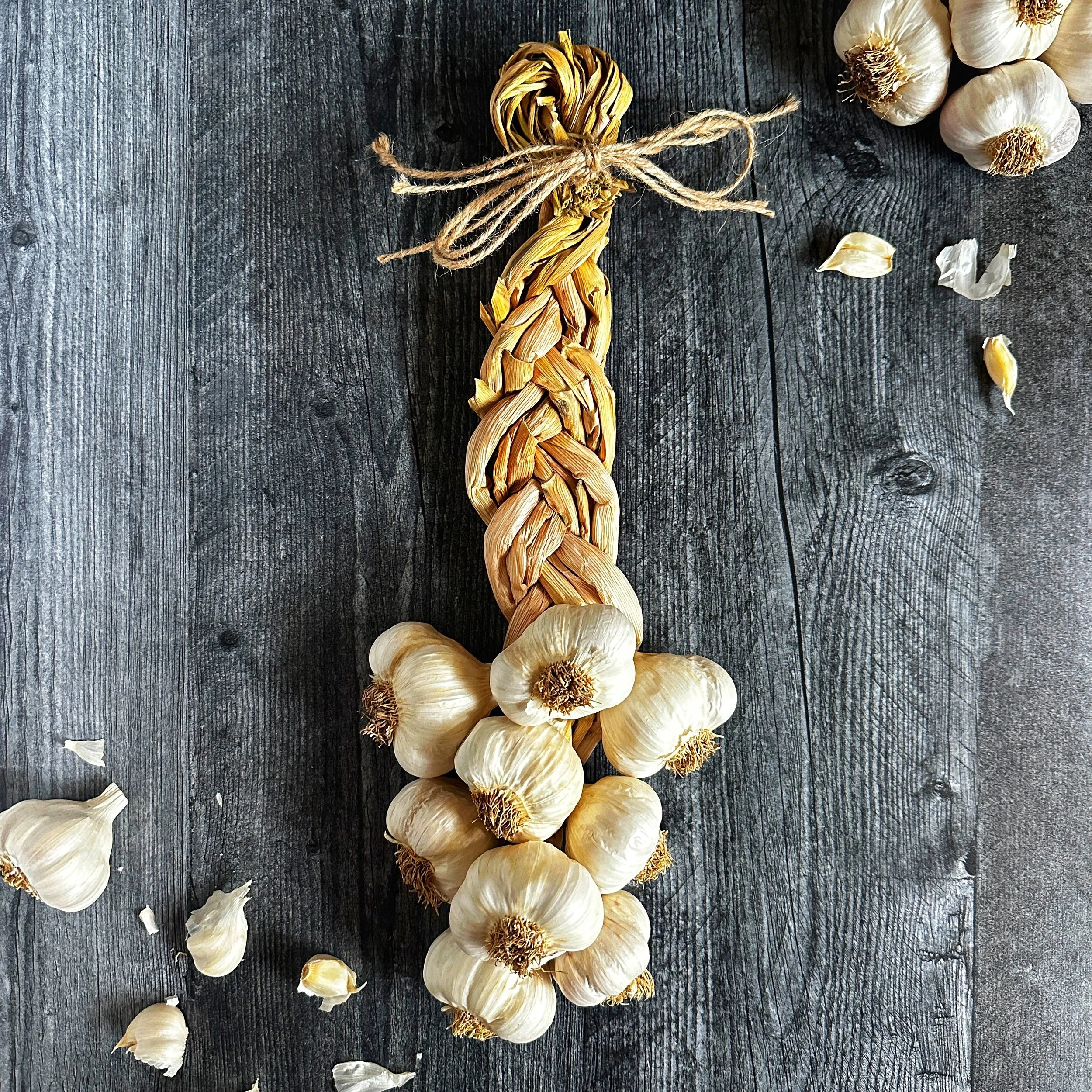 Centered flat-lay photograph of a handmade 9-bulb Mild French garlic braid resting vertically against a dark wood background.