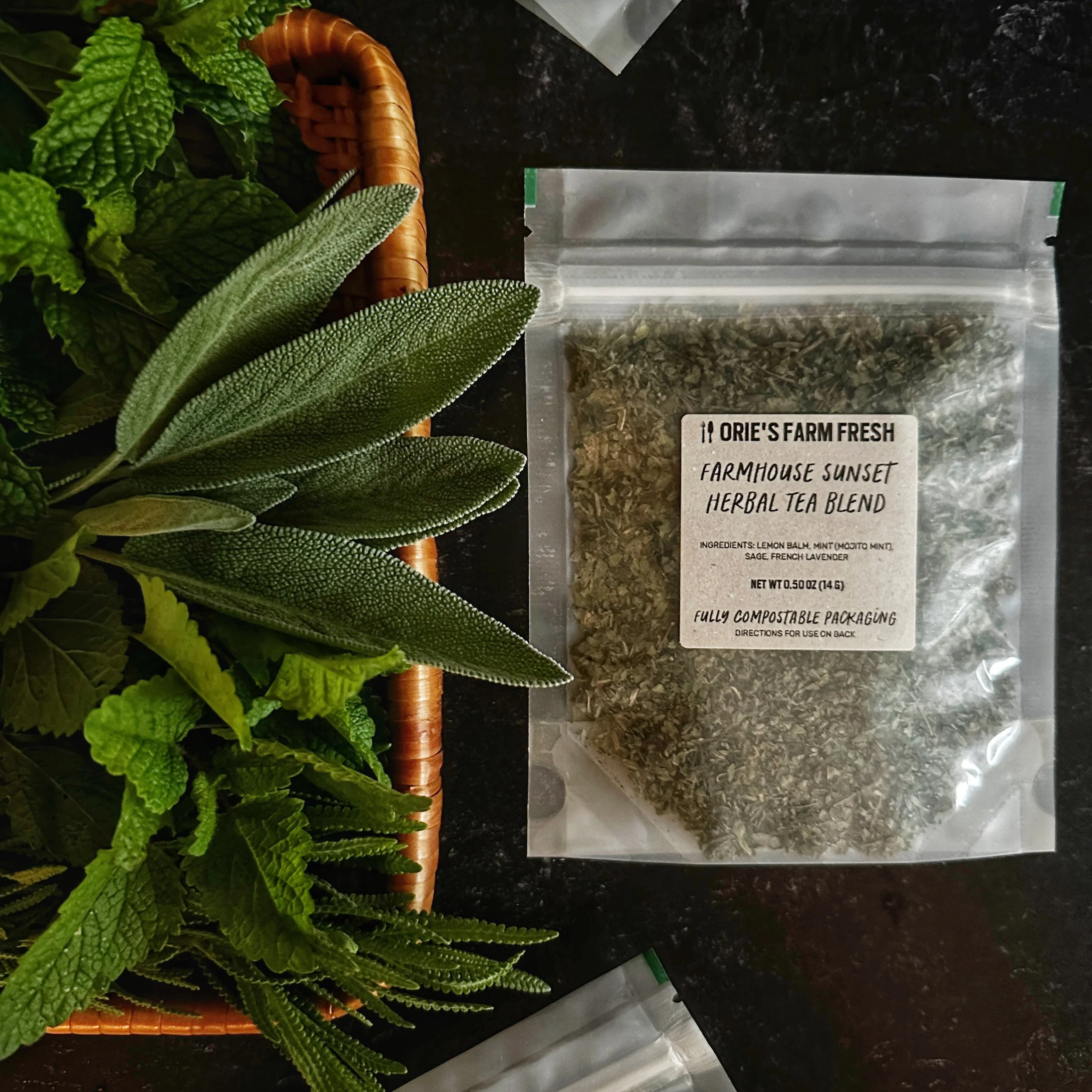 A flat-lay shot of Farmhouse Sunset Herbal Tea Blend  zoomed in to show one package of tea alongside a display of fresh herbs, denoting the tea contains lemon balm, mint, sage and French lavender.