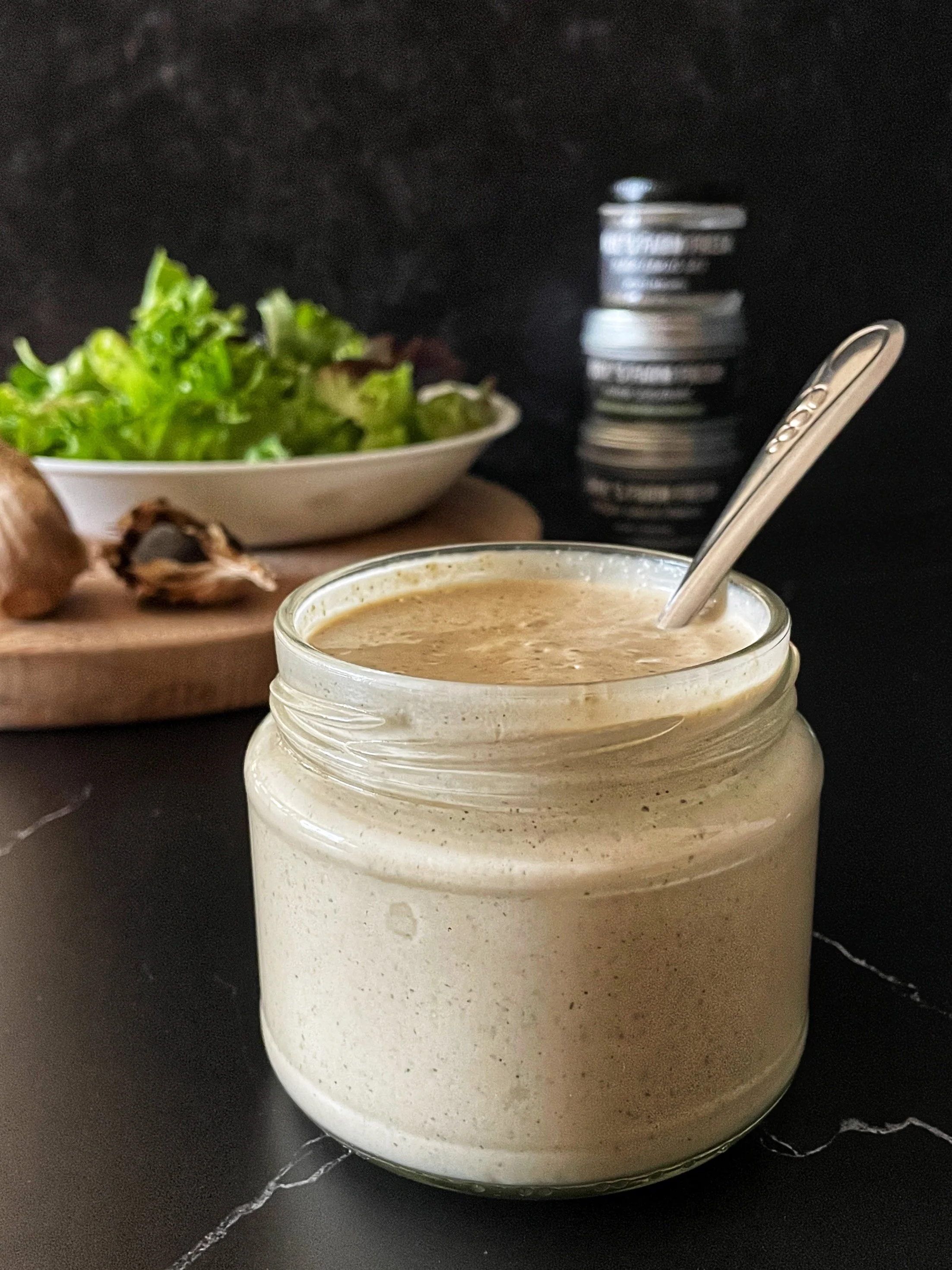 A freshly made jar of Black Garlic Buttermilk Ranch sits in the foreground waiting to be added to a farm fresh salad situated on a wooden cutting board in the background.