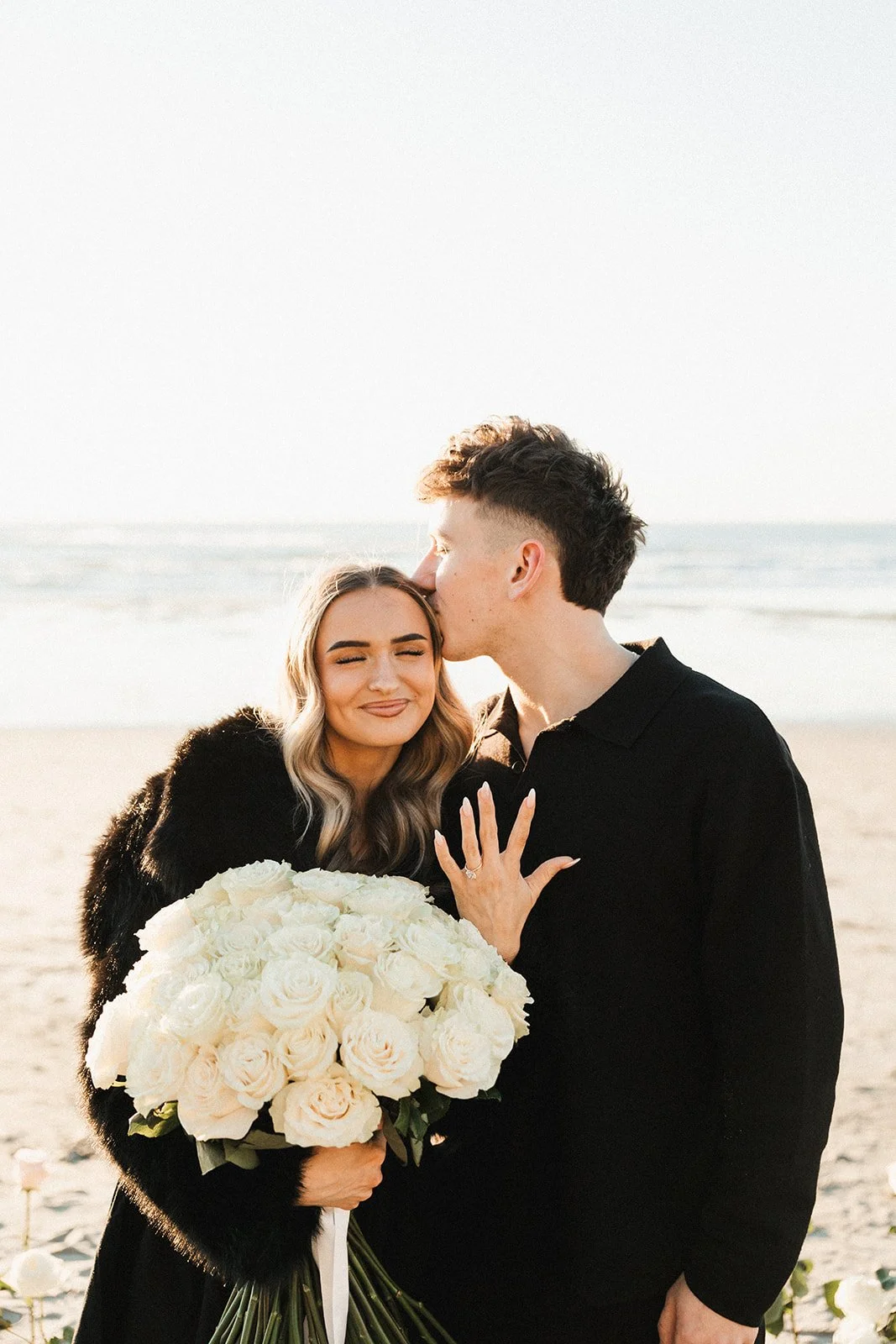 A couple on the beach, with the man kissing the woman's forehead while she holds a large bouquet of white roses. The woman is smiling with her eyes closed, and the man is dressed in black. The background shows the ocean and a clear sky.