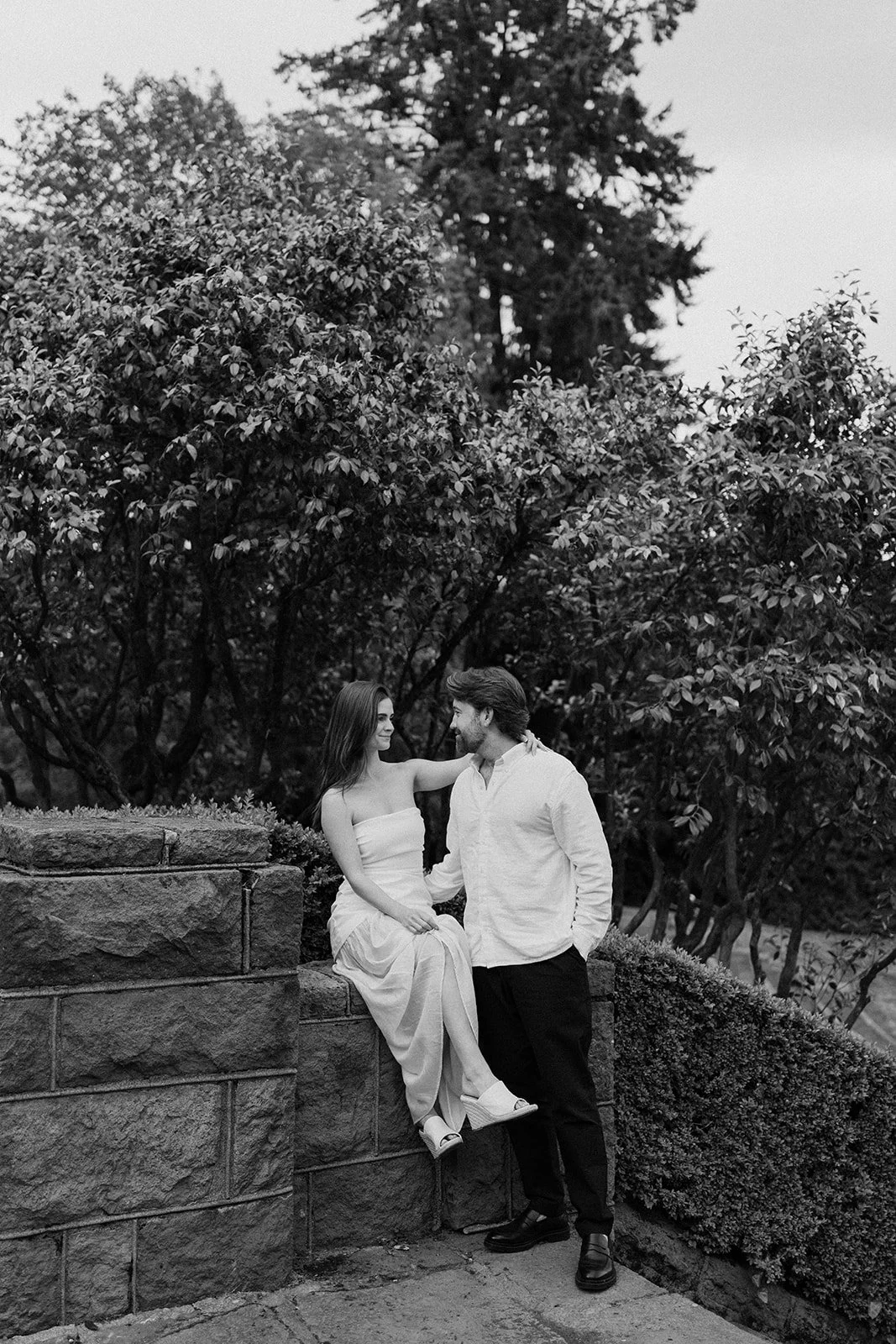 A black and white photo of a couple outdoors, with the woman sitting on a stone wall and the man standing beside her, looking at each other, surrounded by trees and bushes.