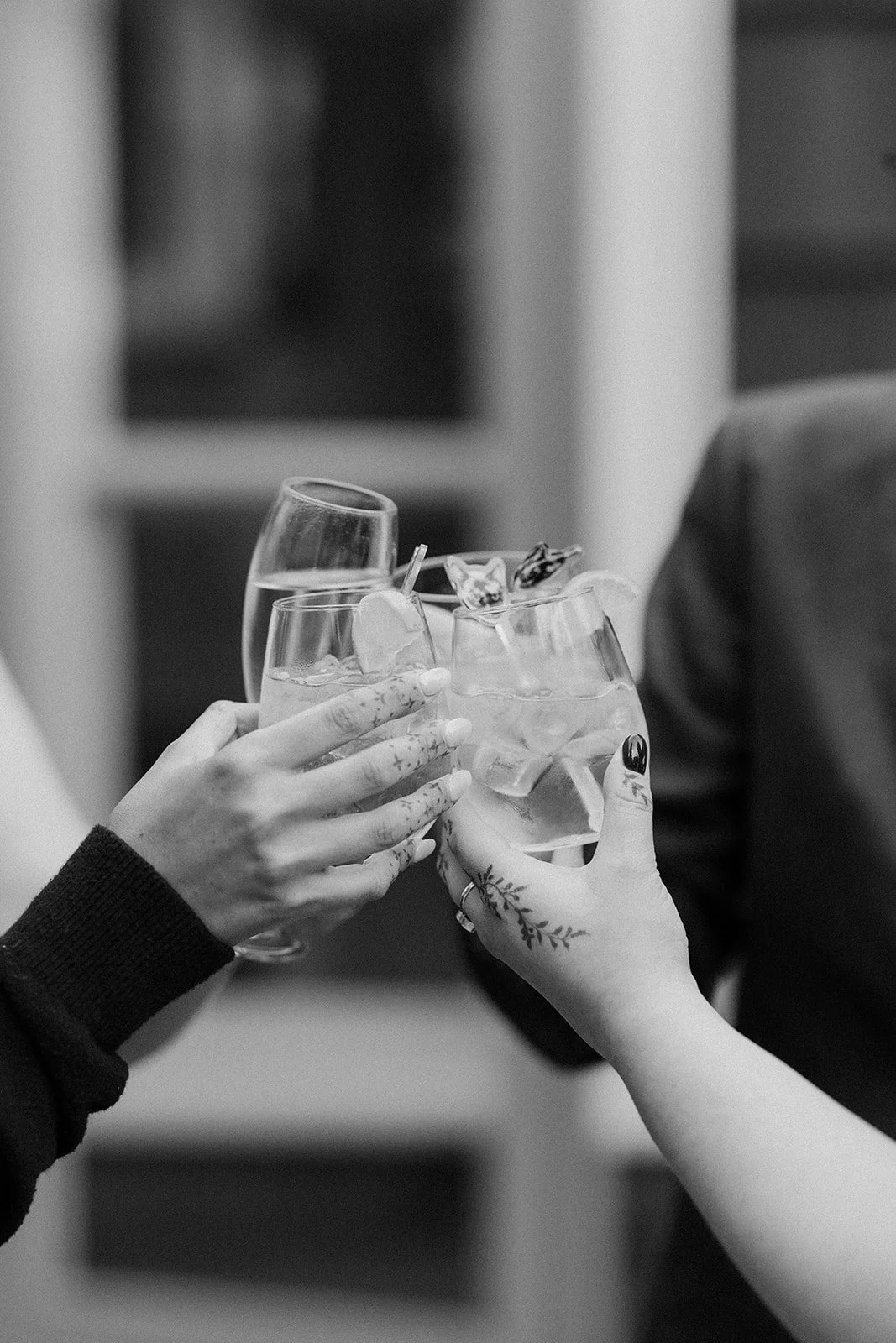 Two people clinking glasses with ice and garnishes in a toast, showing their hands and part of their arms, one hand has floral tattoos.