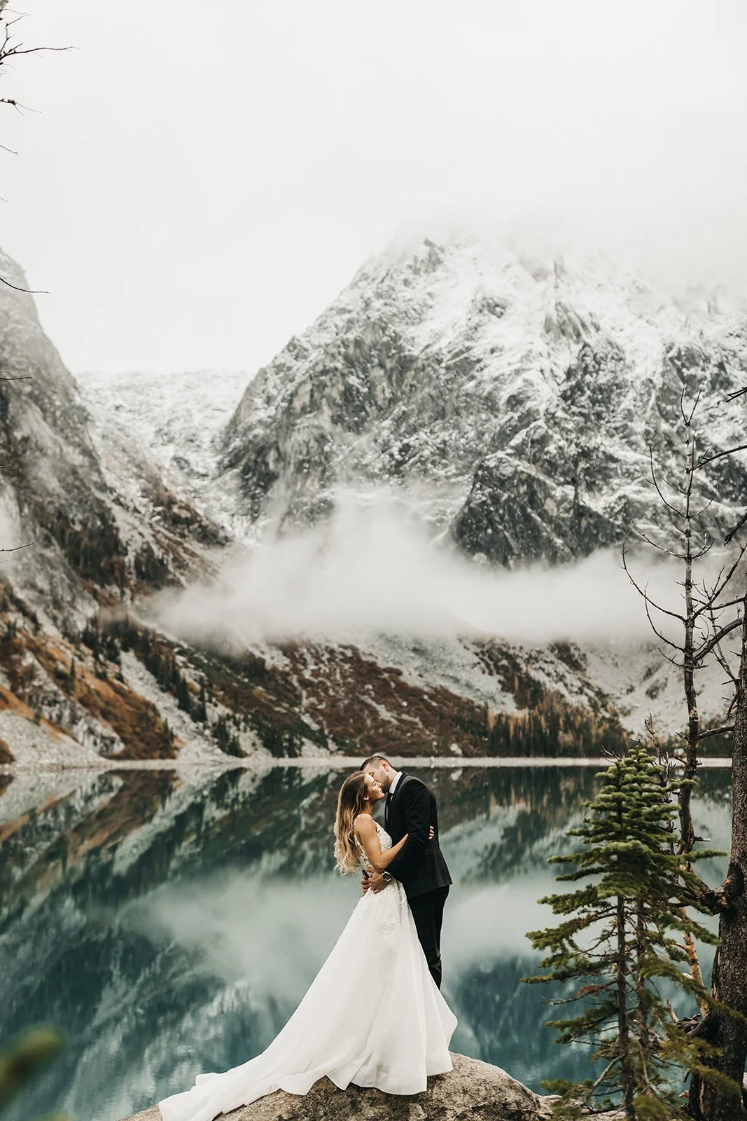 A bride and groom in wedding attire embracing and kissing by a mountain lake, with snow-capped mountains and mist in the background.