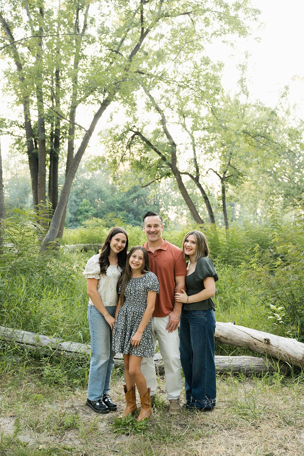 A family of four smiling and standing outdoors in a wooded area with tall trees and green foliage during daytime.