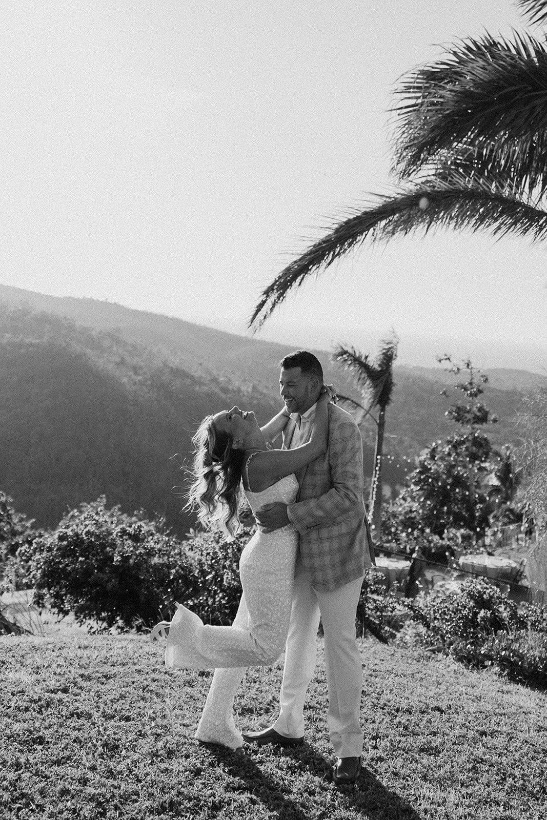 A black and white photo of a couple dancing outdoors, with mountains and palm trees in the background.