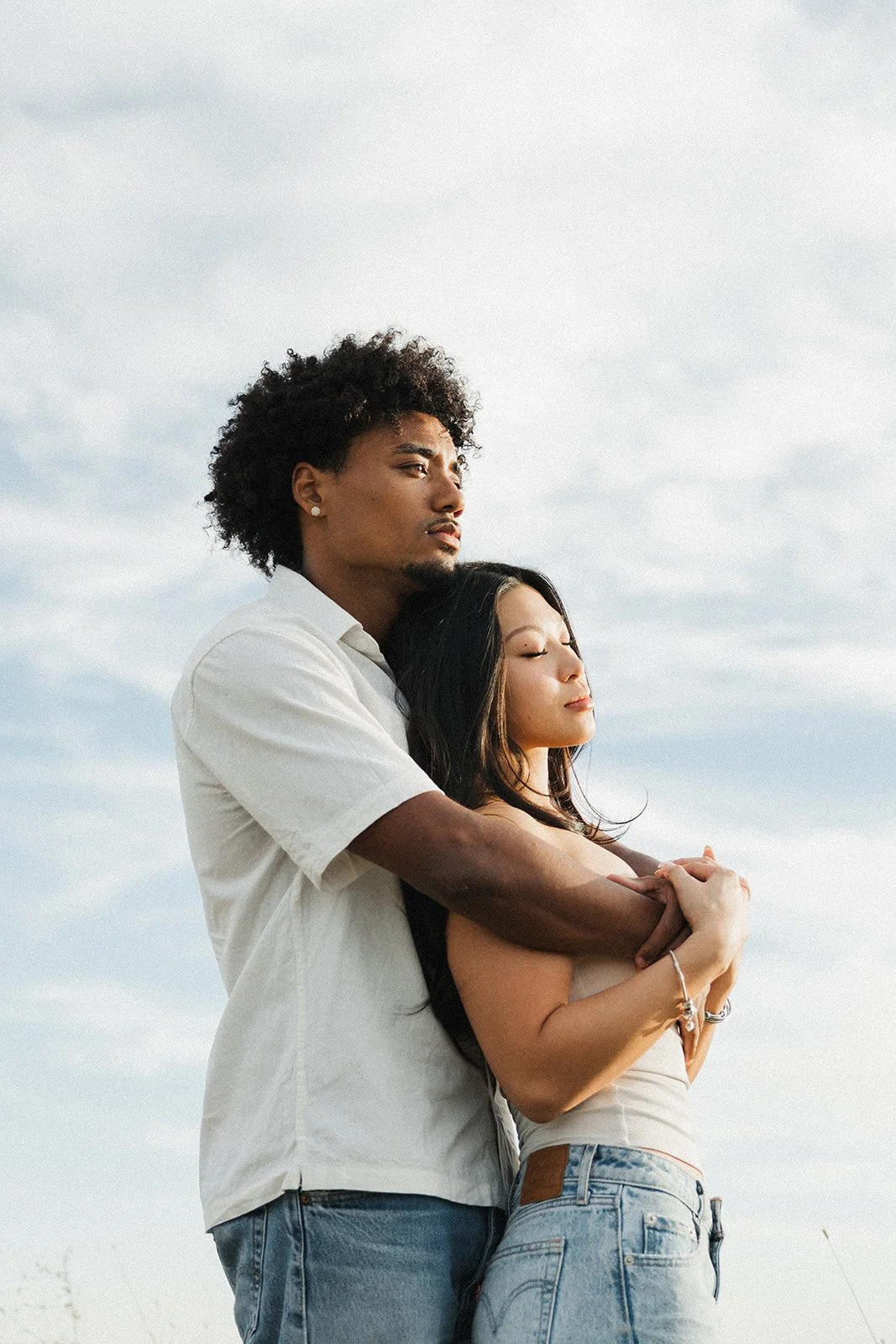 A man and woman embracing outdoors on a cloudy day, with eyes closed and serene expressions.