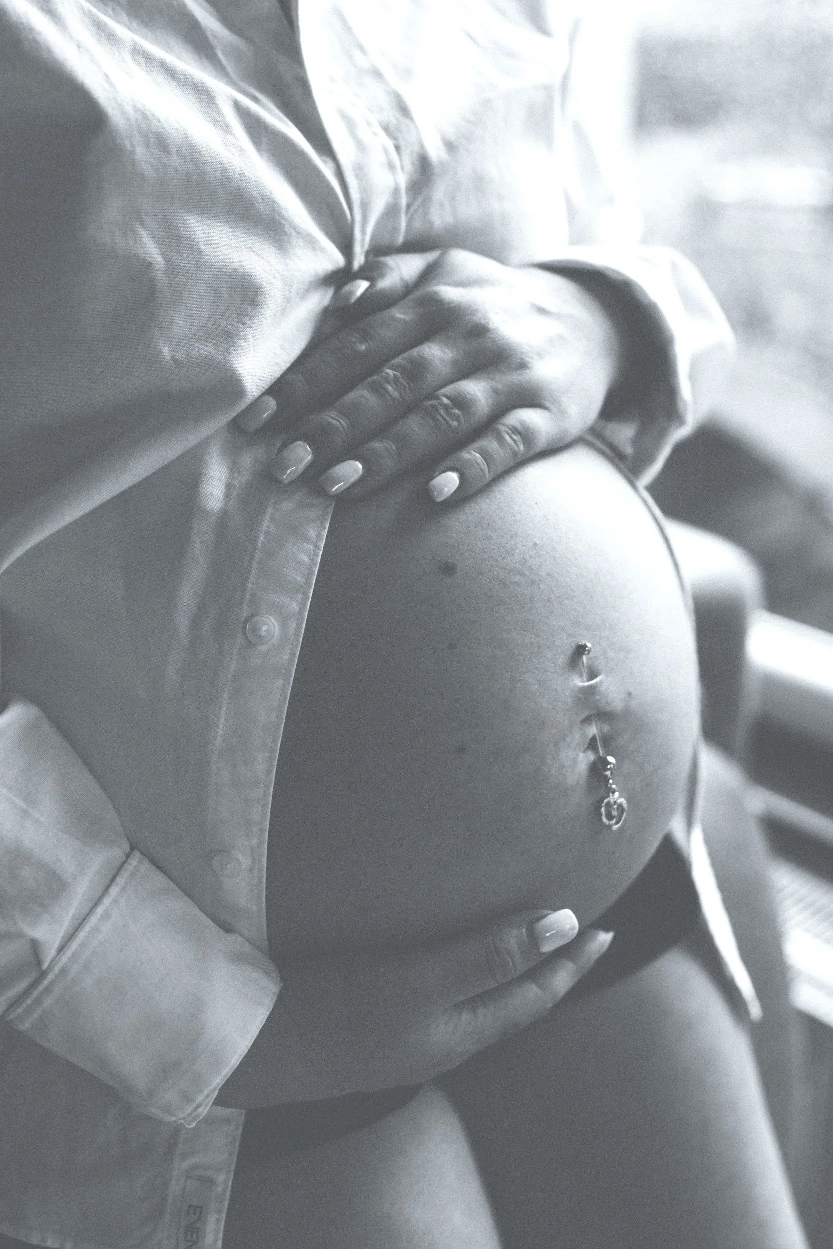 Close-up of a pregnant woman's belly with a belly button piercing, partly covered by her hand and shirt.