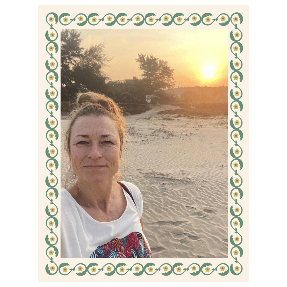 A woman taking a selfie on a sandy beach during sunset, with trees and a few buildings in the background, framed by a decorative floral border.