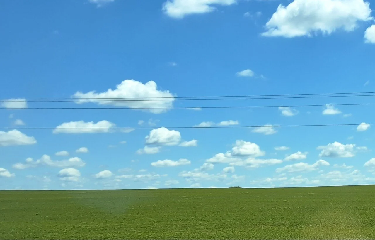 A cultivated landscape stretches toward the horizon beneath an expansive Midwestern sky, where utility lines mark the intersection of agricultural land, infrastructure, and the atmosphere above.