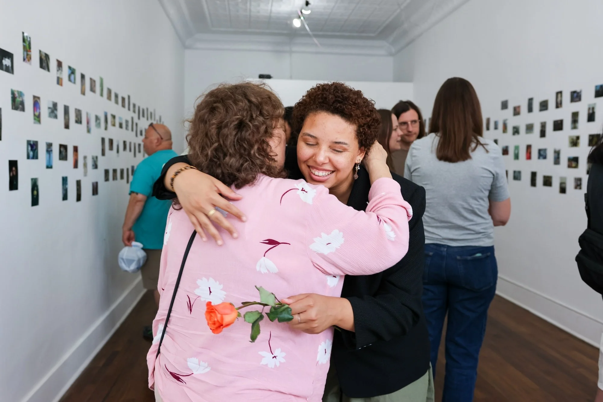 A photograph of two people embracing. One looks towards the camera and smiles warmly while holding a rose. The other, whose back is towards the camera, wears a pink shirt with a floral print.