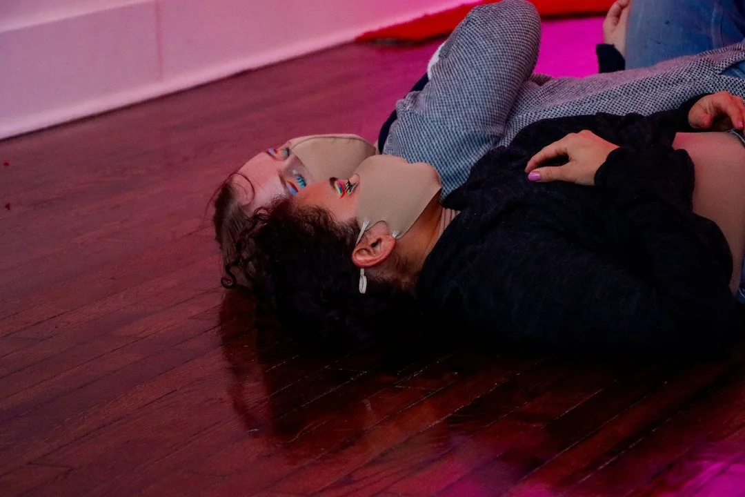 A photograph of two dancers lying close to one another on a dark hardwood floor. Their eyes are closed; they wear face masks and look as though they’ve just expended an incredible amount of energy.