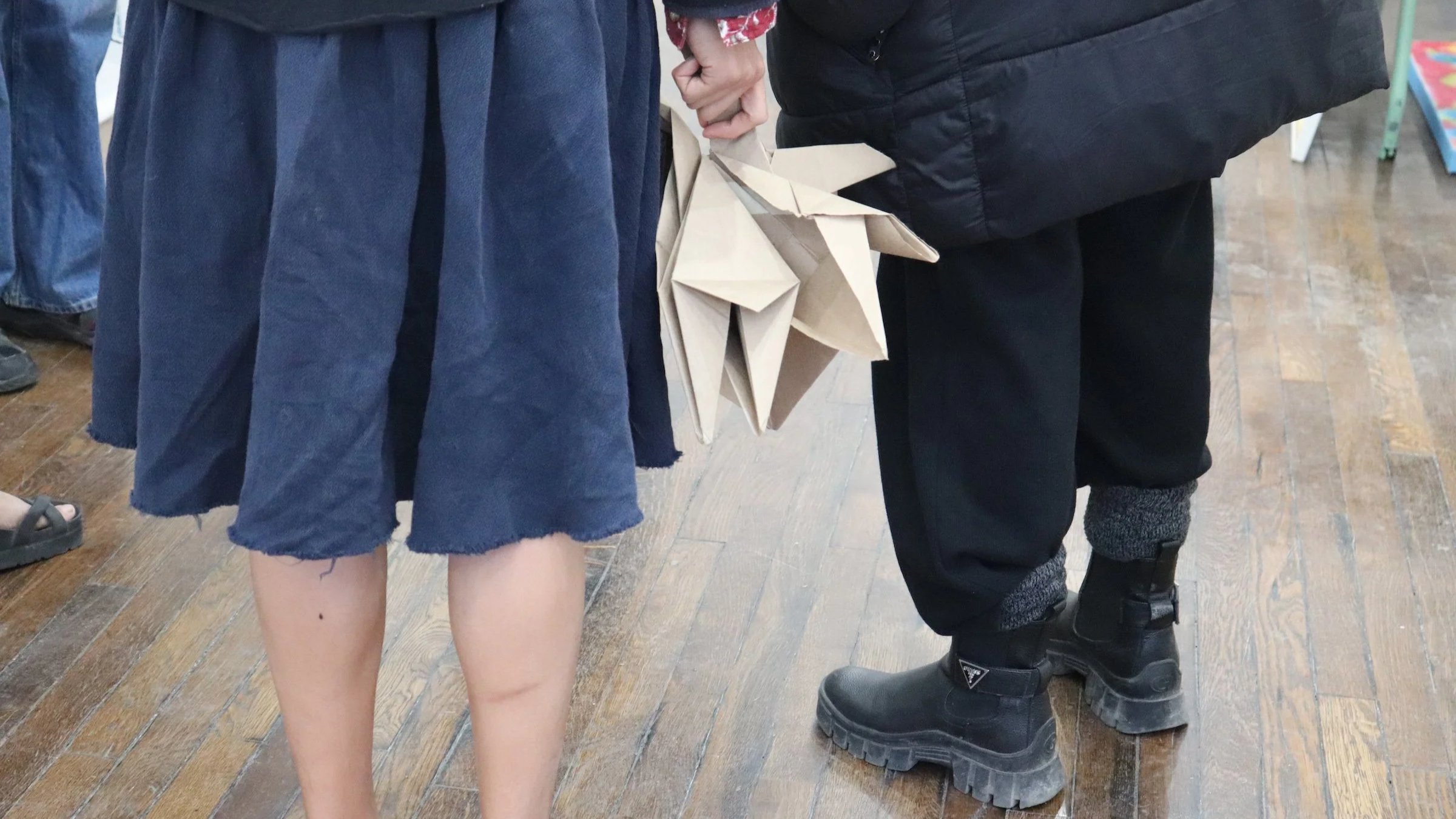 A photograph of an artist holding an elaborately folded work made of brown paper that resembles a large-scale cootie catcher. They hold it casually, letting it dangle at their side.